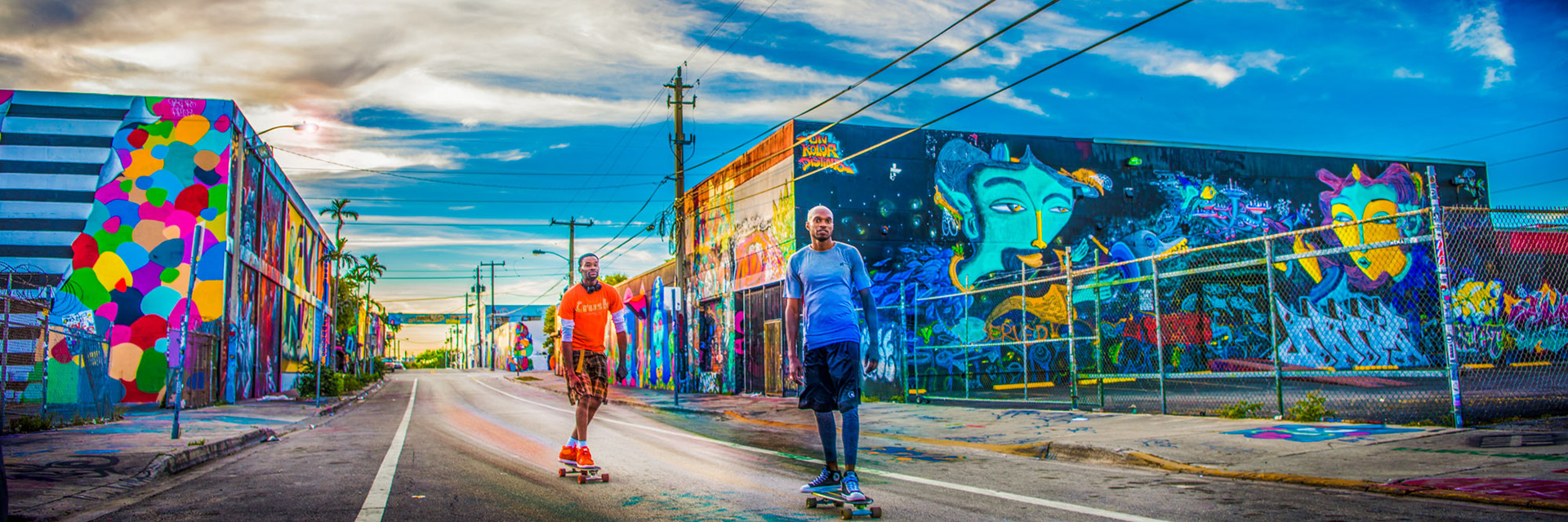 Two people skateboarding on a vibrant city street with colorful murals. The bright sky and artistic atmosphere convey a lively, urban vibe.