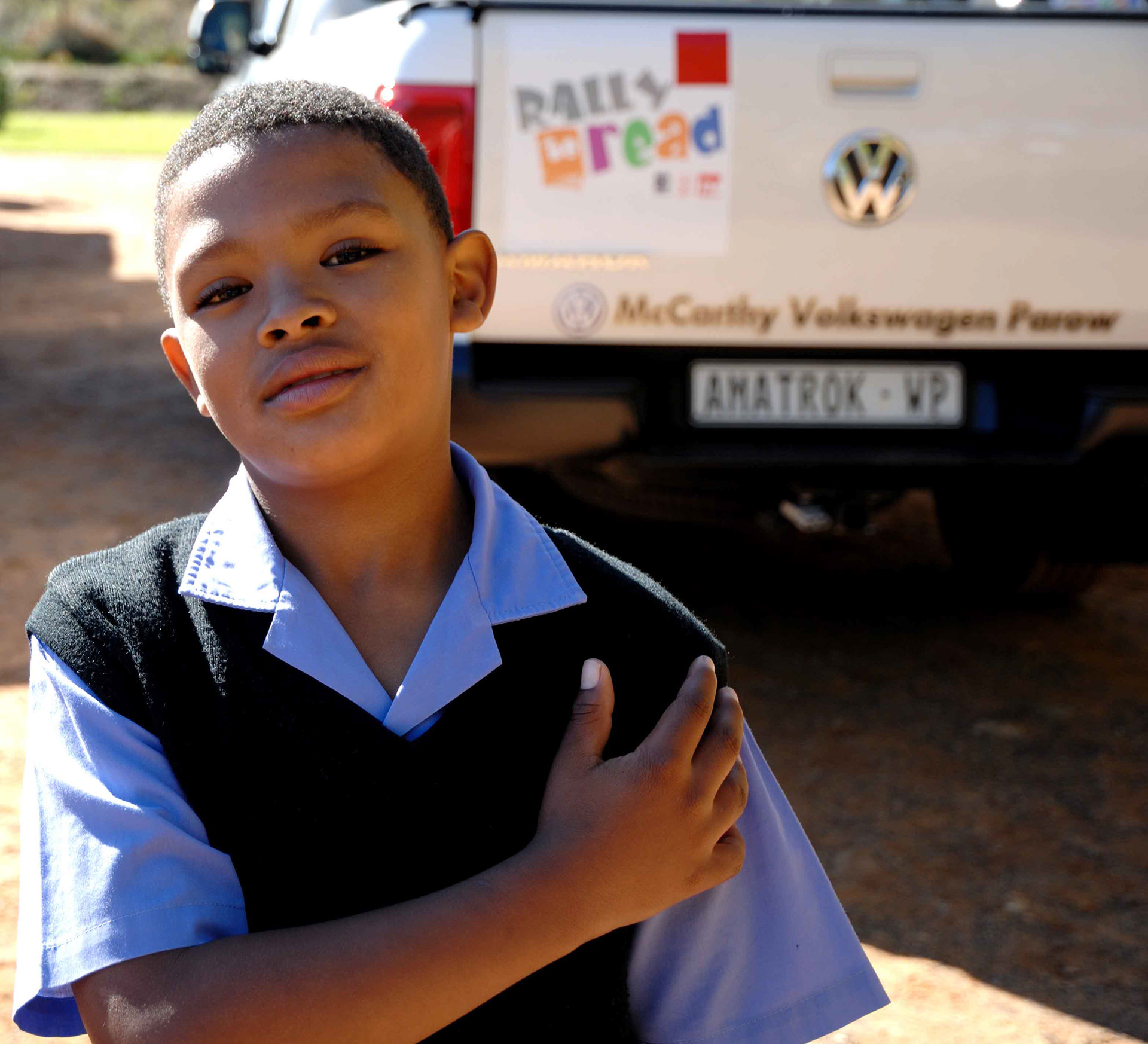 A young person stands confidently, wearing a blue shirt and black vest. In the background, a white Volkswagen with "Rally to Read" is visible, suggesting a community event.