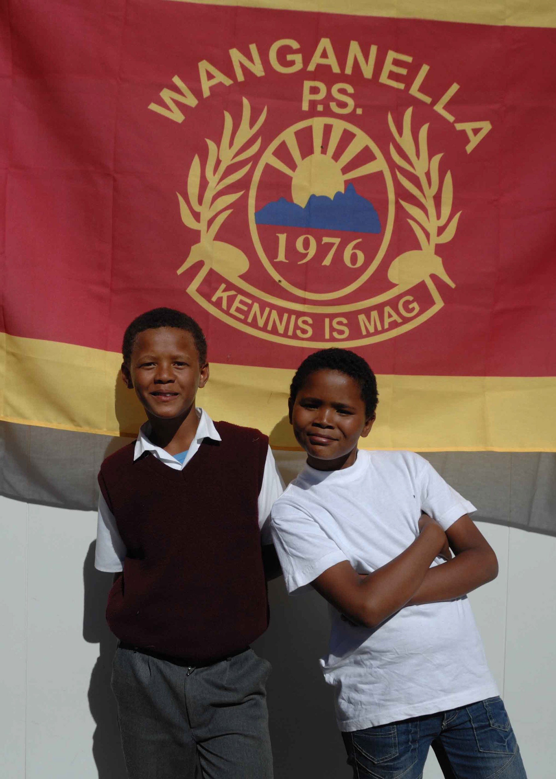 Two children smiling in front of a red and yellow banner with "Kennis is Mag" and "1976." The banner has a laurel wreath and sun design.