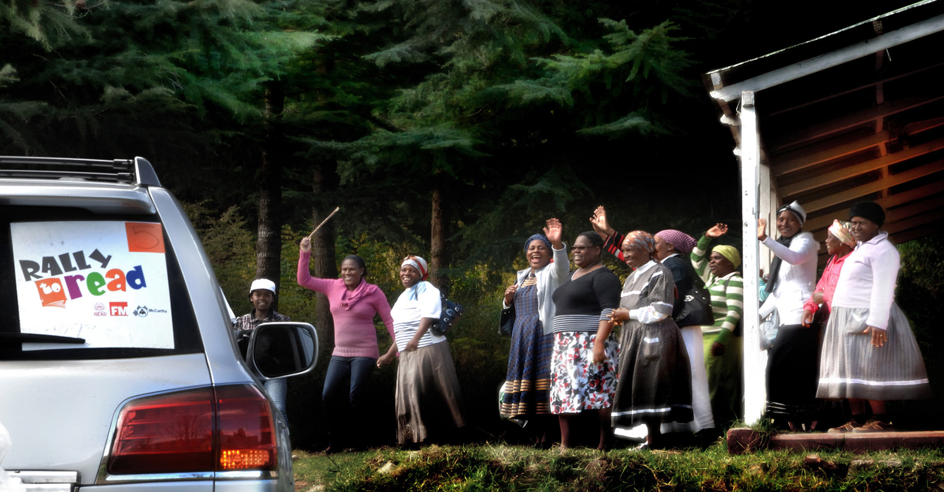 A group of smiling people waving next to a building, with a car labeled "Rally to Read" in the foreground. The scene is set in a wooded area.