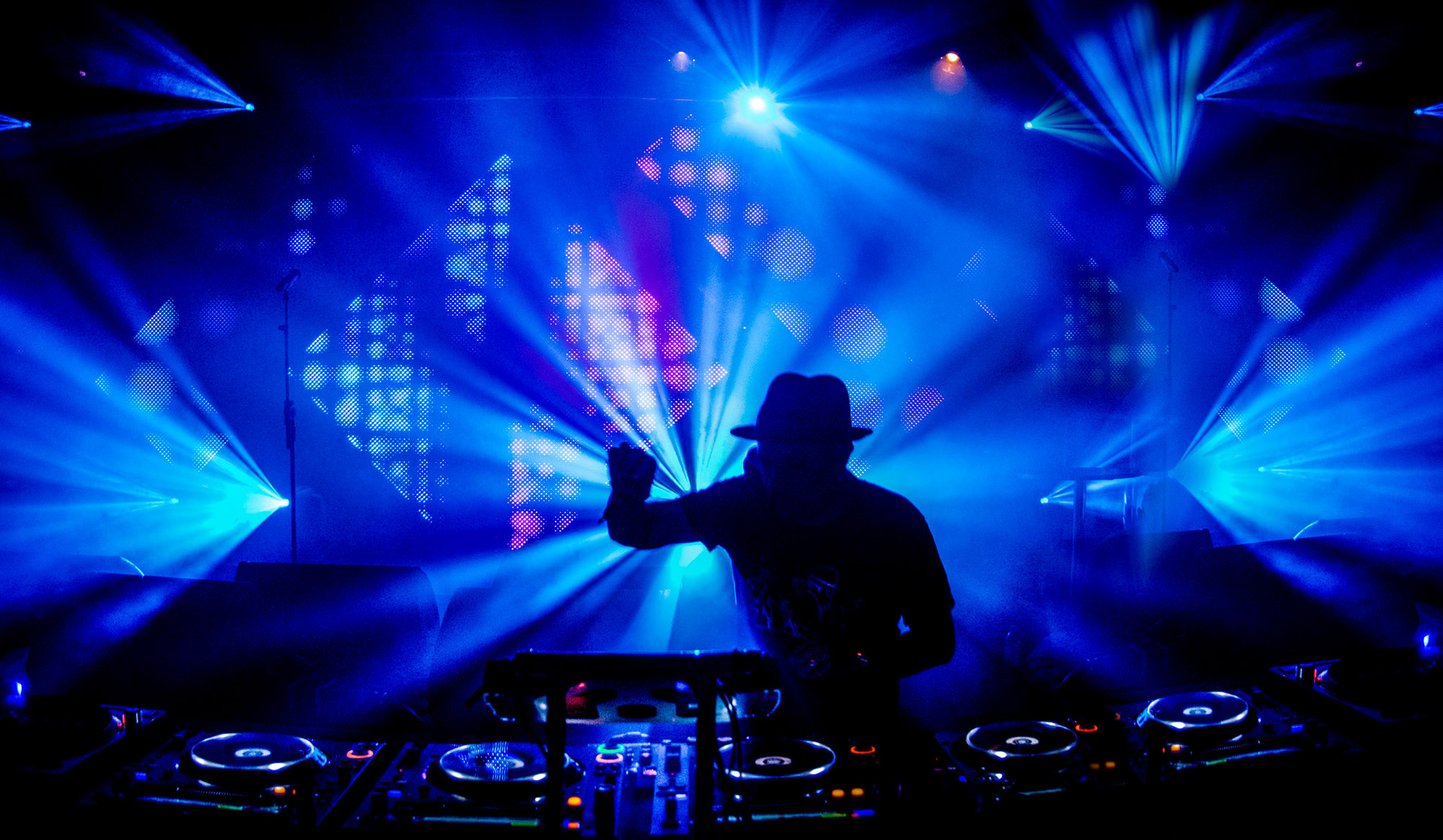 Louie Vega DJ silhouette against vibrant blue and purple stage lights, standing at a mixer, creating an energetic, dynamic atmosphere in a concert setting.
