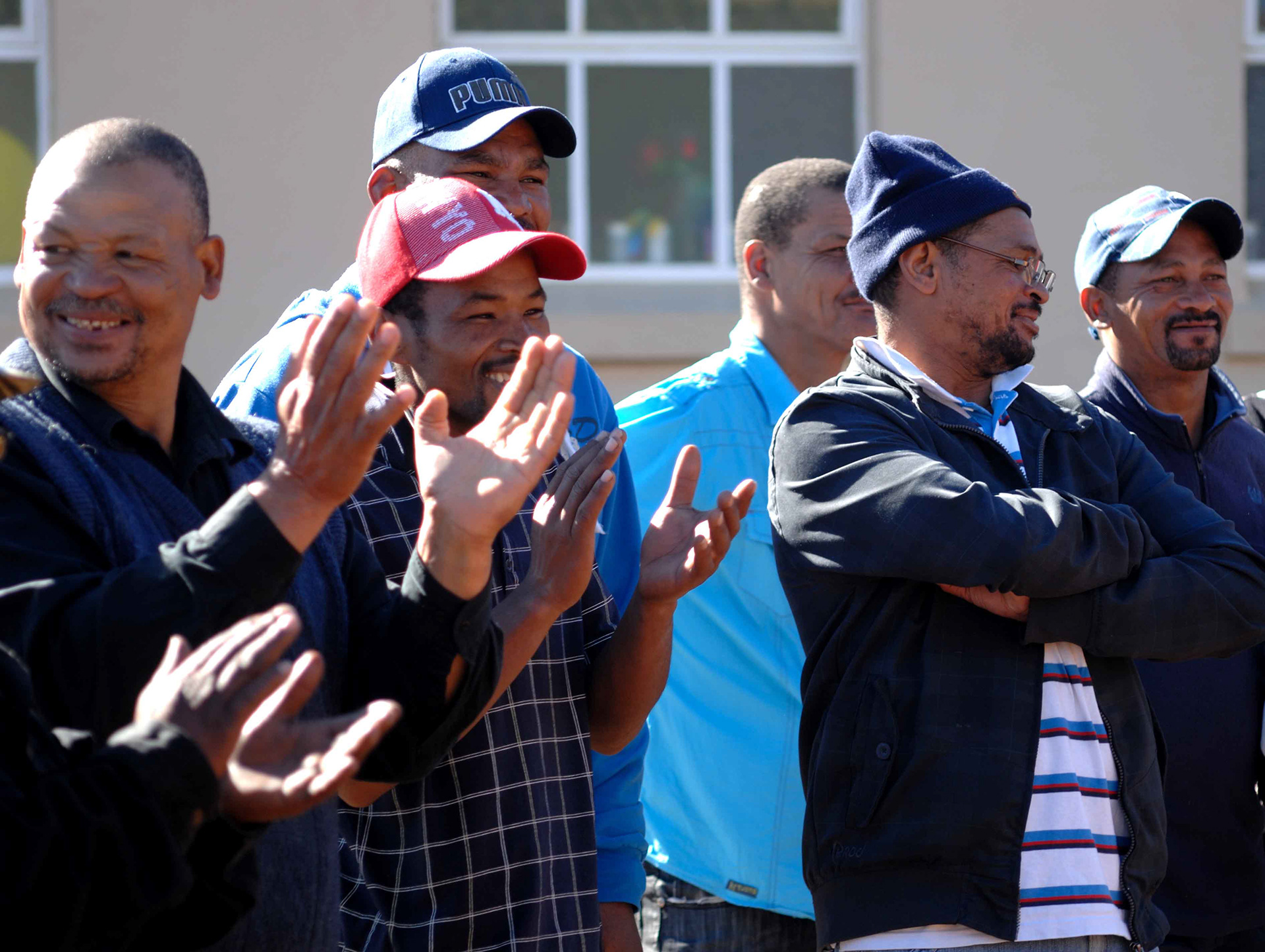 A group of men, wearing caps and smiling, clap enthusiastically outdoors. Their expressions convey happiness and community spirit in a sunny setting.