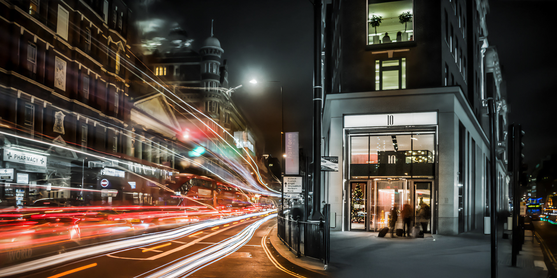 Night-time city street scene with blurred light trails from passing cars, highlighting motion. A tall, lit building entrance on the right. A dynamic and vibrant urban atmosphere.