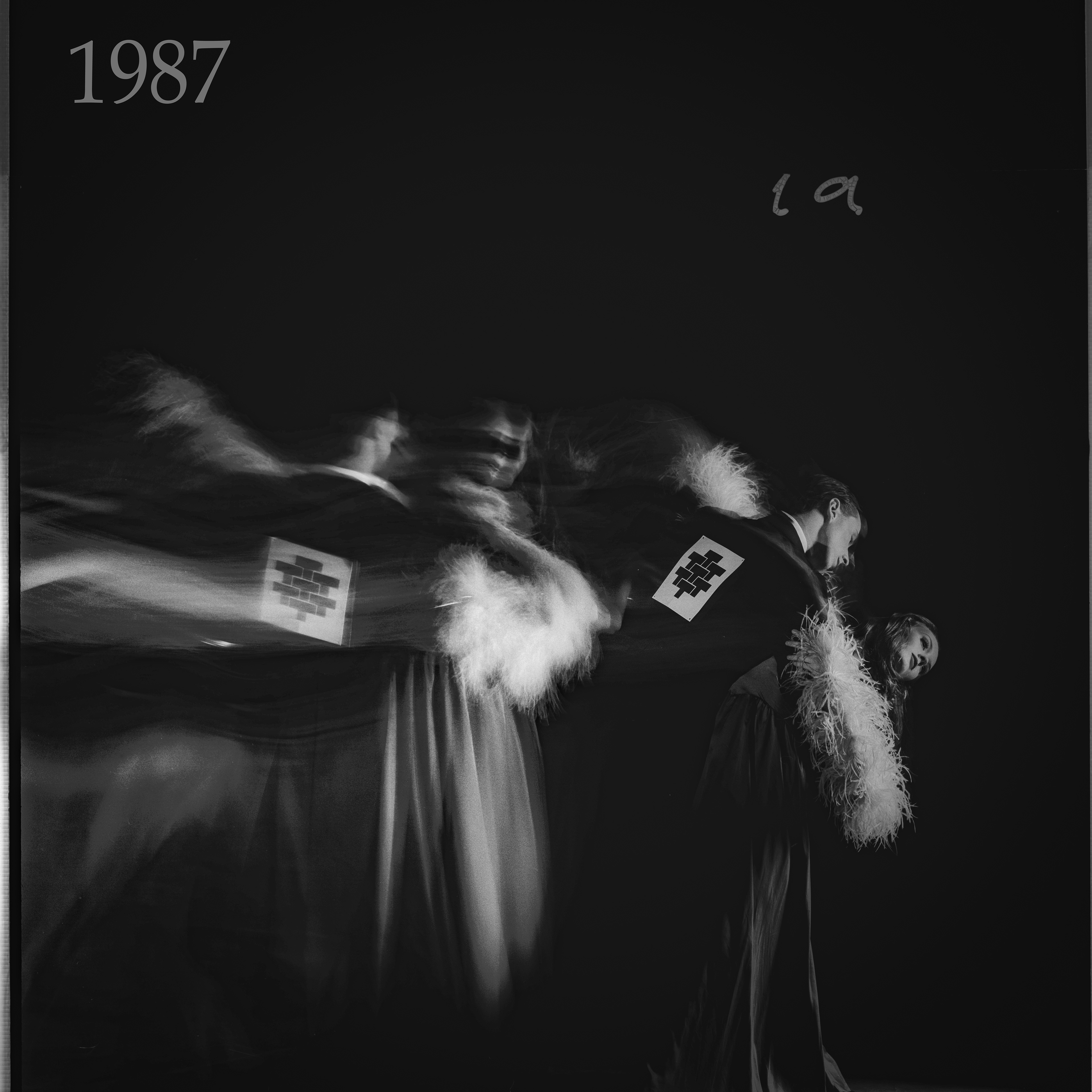 Blurred black and white image of dancers with feathered costumes and numbered armbands. The number "1987" is seen, adding a nostalgic feel.