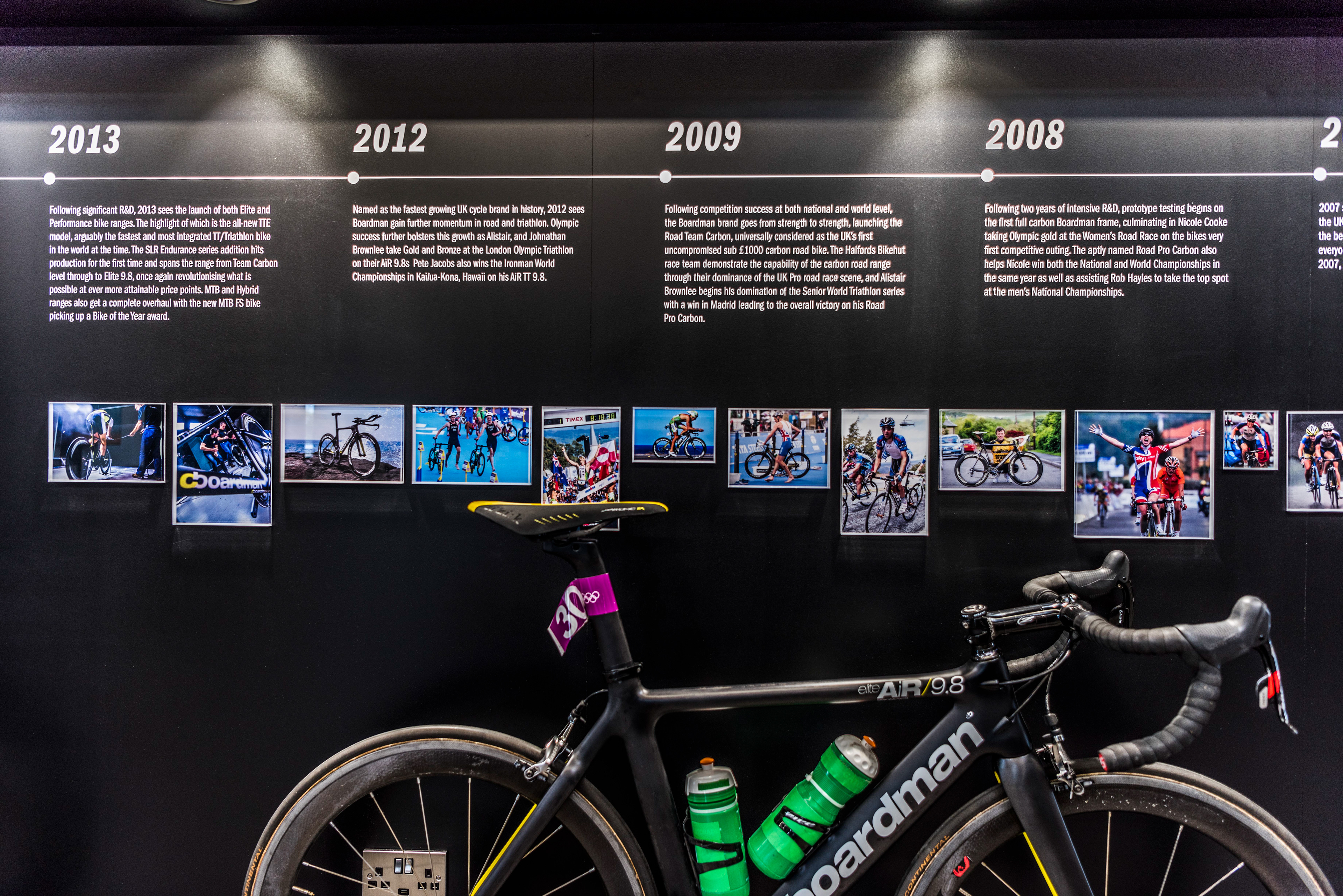 Exhibit timeline showcases bicycle evolution from 2008 to 2013. Photos of cyclists accompany text. A black Boardman bike with green water bottles is in the foreground.