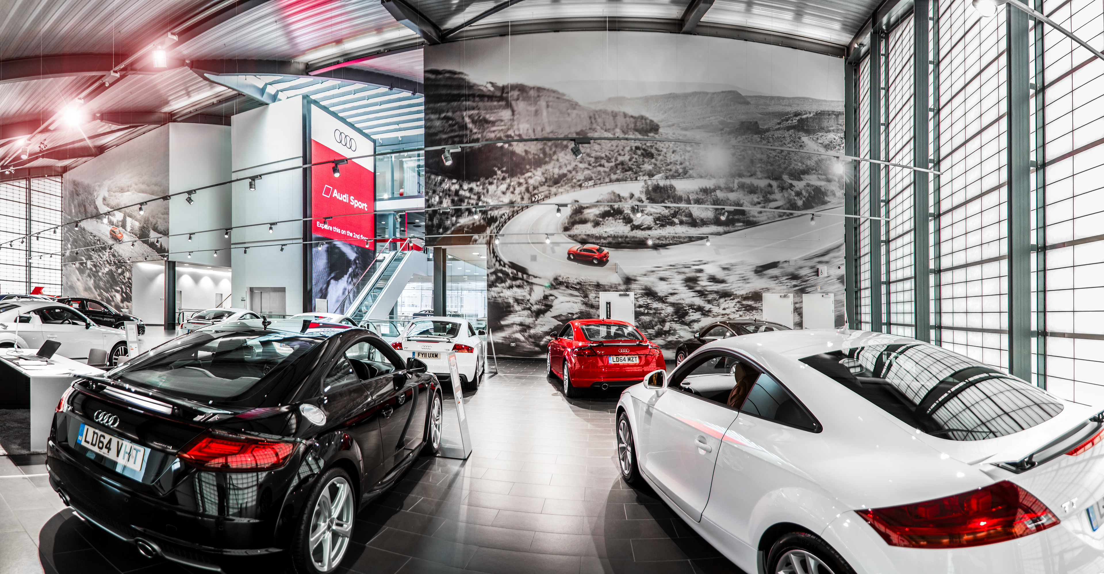 Showroom displaying luxury cars with a red and black Audi in the foreground. Large mural depicts a dynamic car on a winding road. Modern, sleek atmosphere.