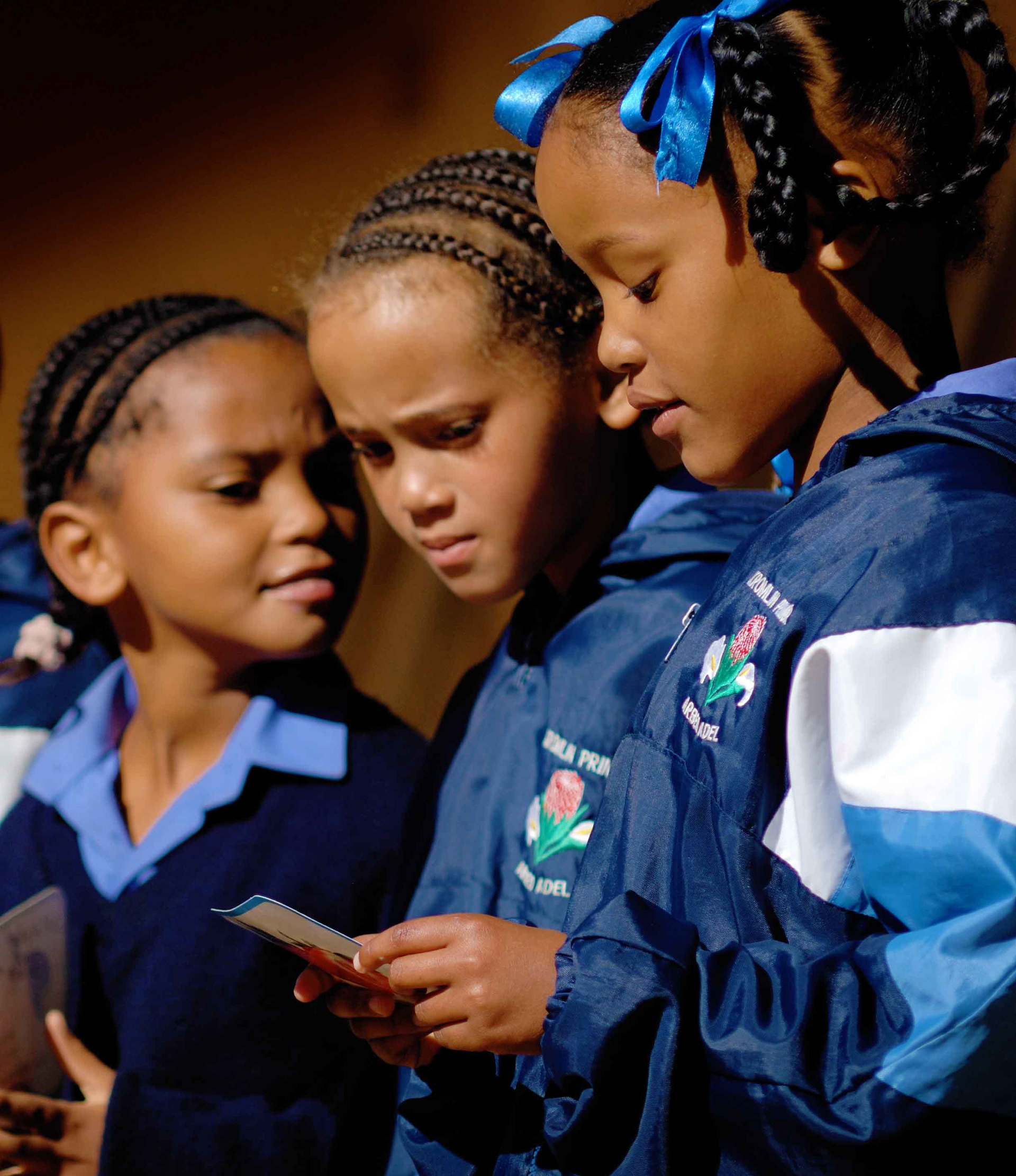 Three young girls with braided hair in blue outfits and bright blue bows stand closely together. They display focused expressions in soft lighting.