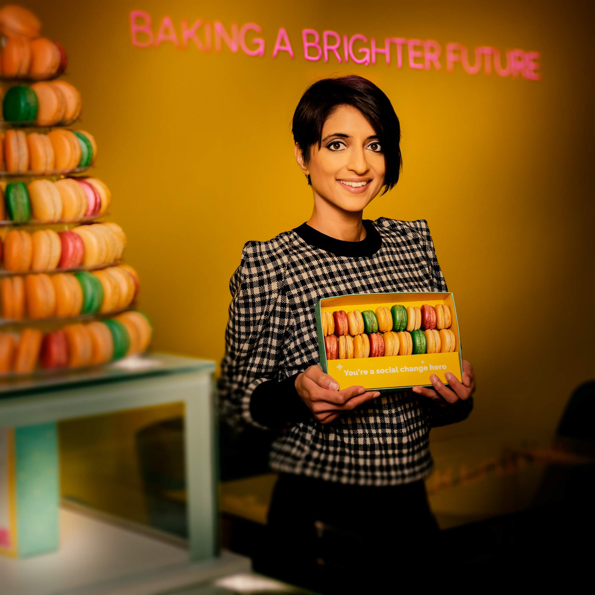 A person smiles while holding a box of colourful macarons with the text "You're a social change hero." Background displays more macarons, creating a cheerful, vibrant scene.