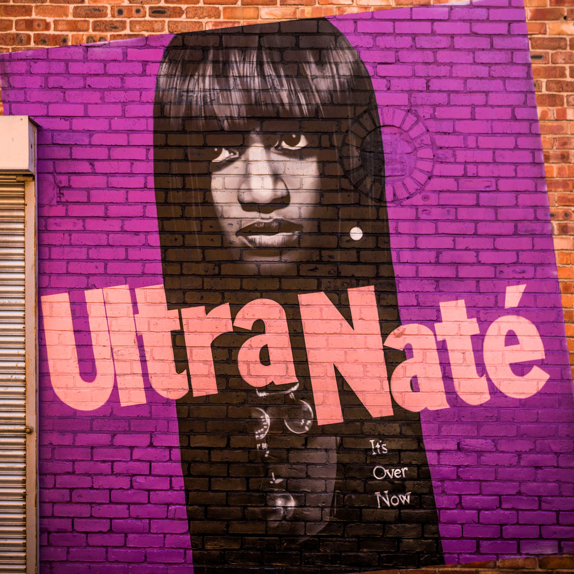 Street art mural on a brick wall features a monochrome portrait of a serious-looking woman with text "Ultra Naté" in large pink letters on a purple background.