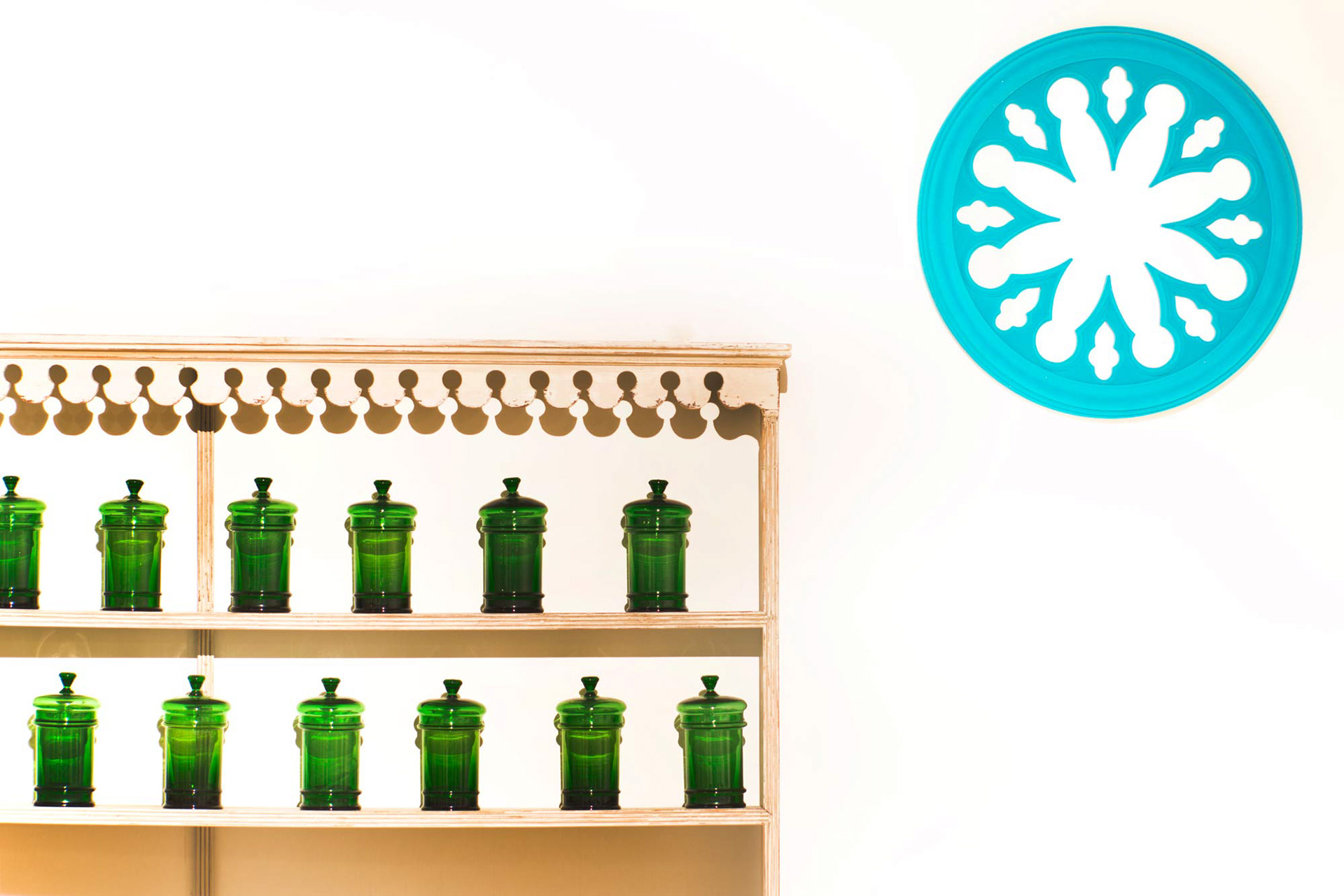 Wooden shelf with green glass jars neatly arranged against a white wall. A blue decorative circular pattern hangs on the right, creating a calm, vintage vibe.