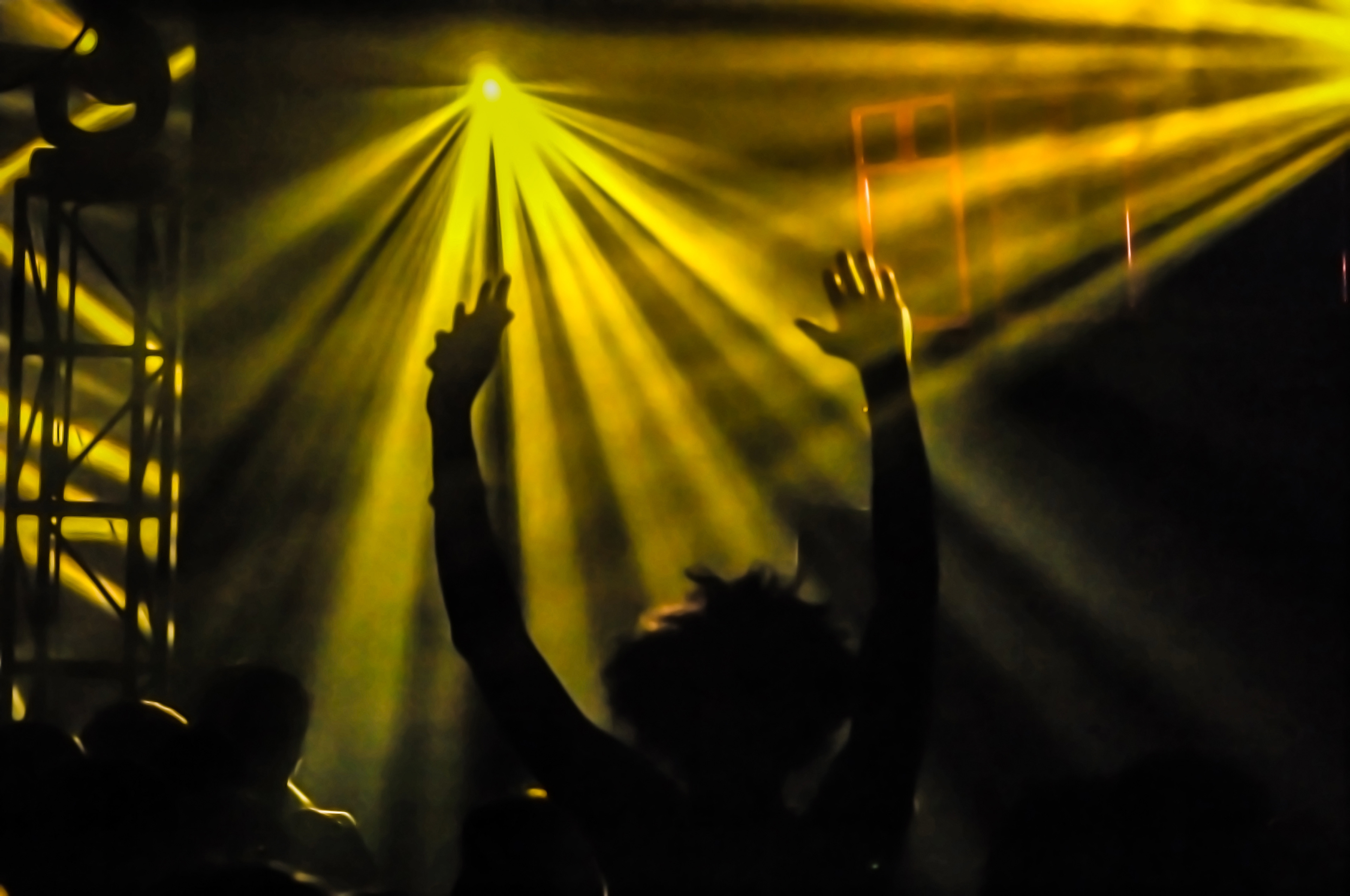 Silhouetted person with raised arms at a concert, illuminated by vibrant yellow stage lights. The atmosphere is energetic and lively.