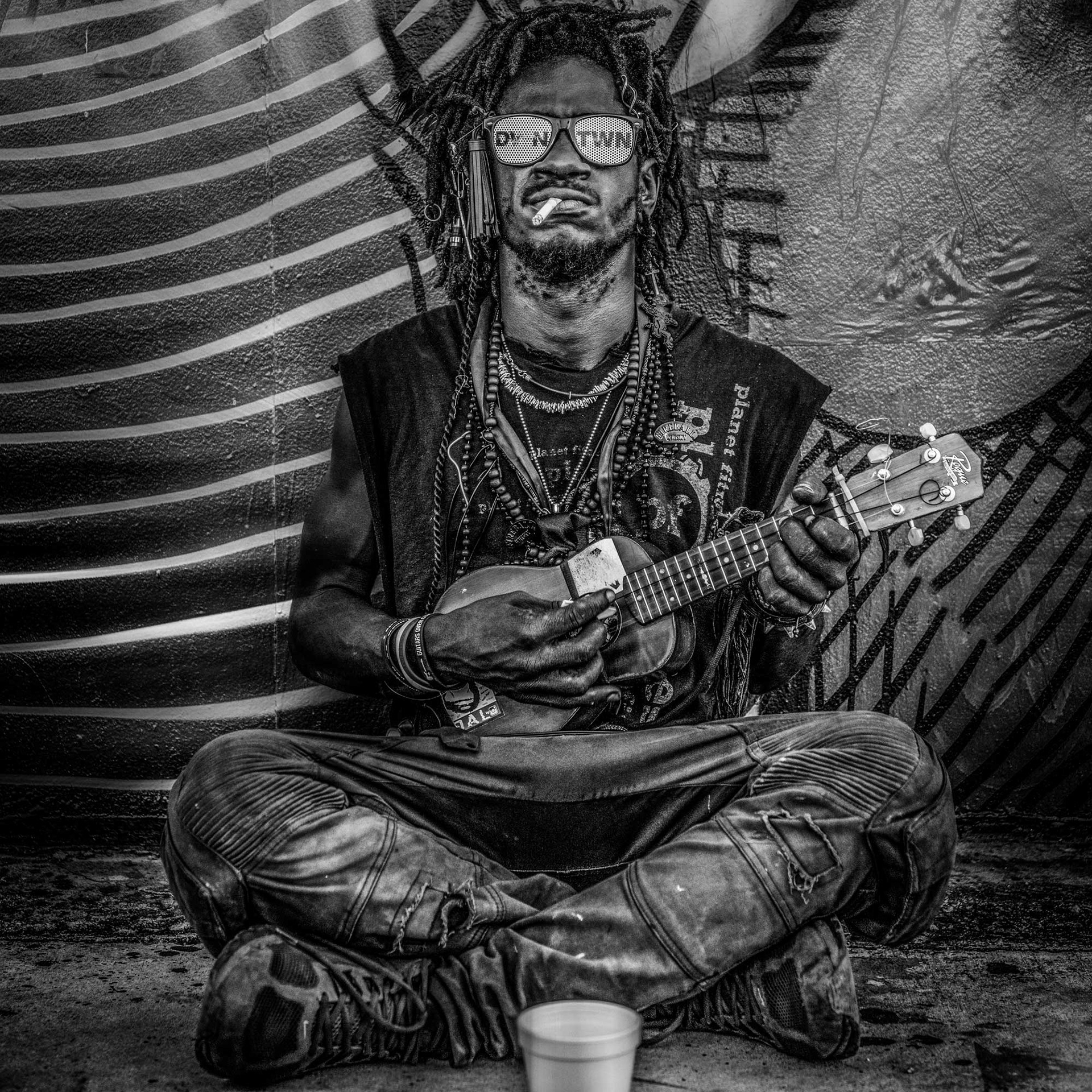 Black and white image of a person with dreadlocks, sunglasses, and layered necklaces playing a ukulele. They sit cross-legged, exuding a relaxed vibe.