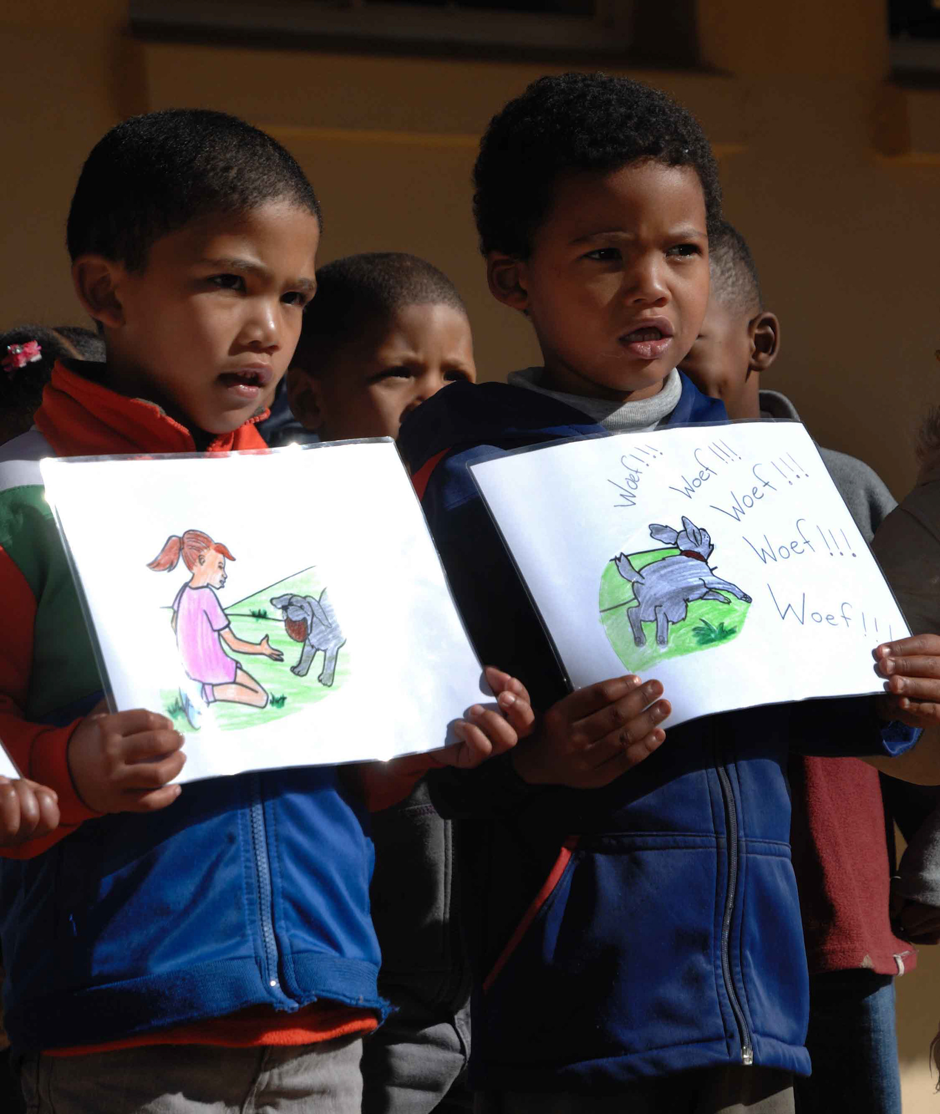 Two young boys hold illustrated sheets. One shows a girl with a dog, the other a barking dog with "Woef!!!" text. Both appear focused and intent.