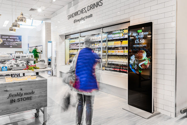 A supermarket interior with a white-tiled wall, a drinks fridge, and product shelves. A digital screen advertises a £5.50 crispy chicken burger, while a person in motion wears a blue vest, creating a sense of energy and bustling activity.
