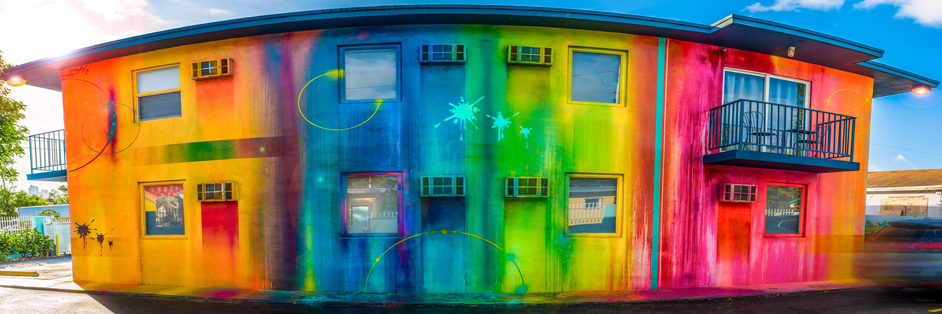 Two-story building with vibrant rainbow graffiti, featuring large splashes of color and circles. Balconies and windows add contrast against a bright blue sky.