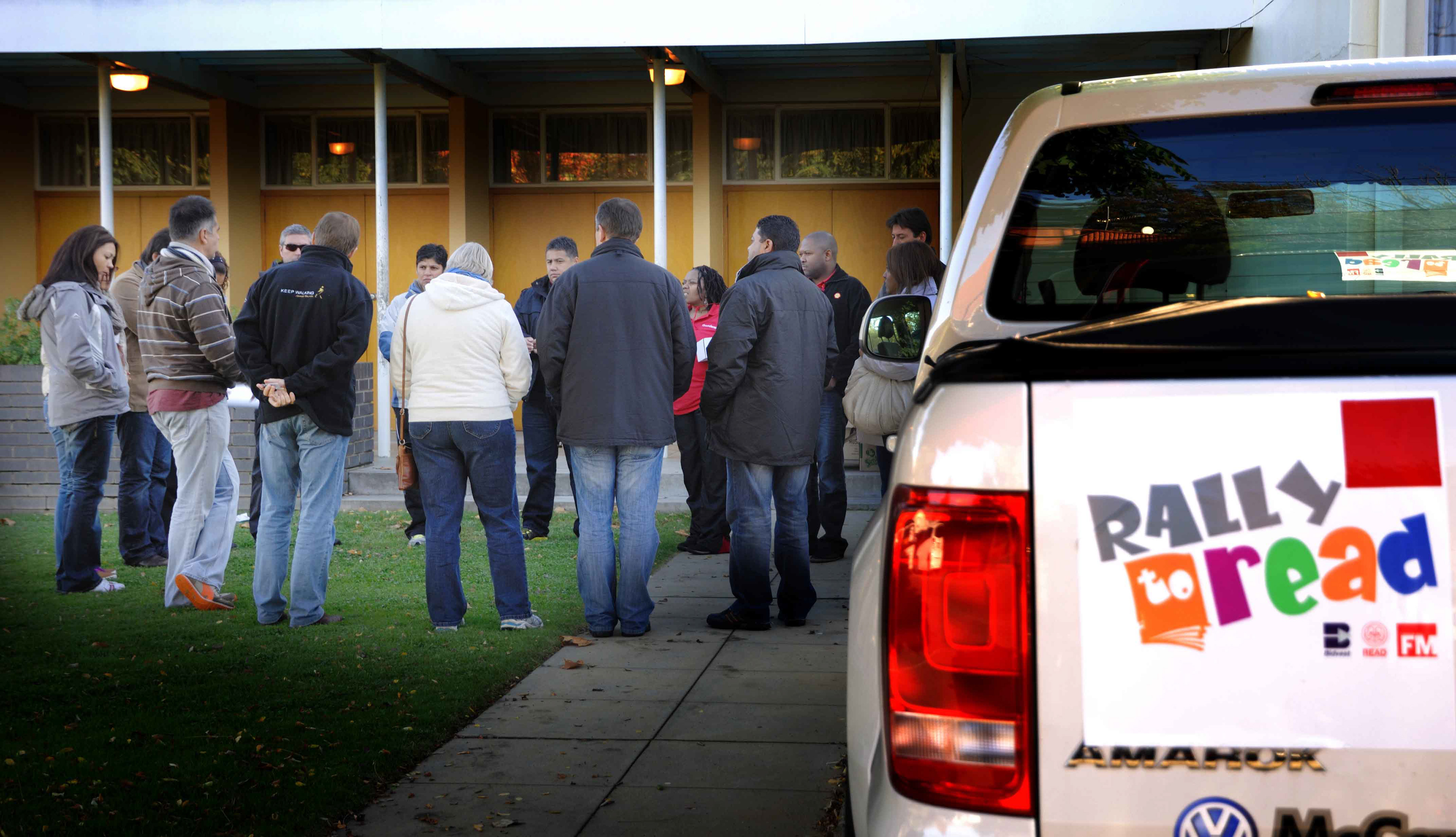 A group of people gathers outside, some with hands in pockets, dressed warmly. Nearby, a vehicle displays a colorful "Rally to Read" sign, suggesting a community event.