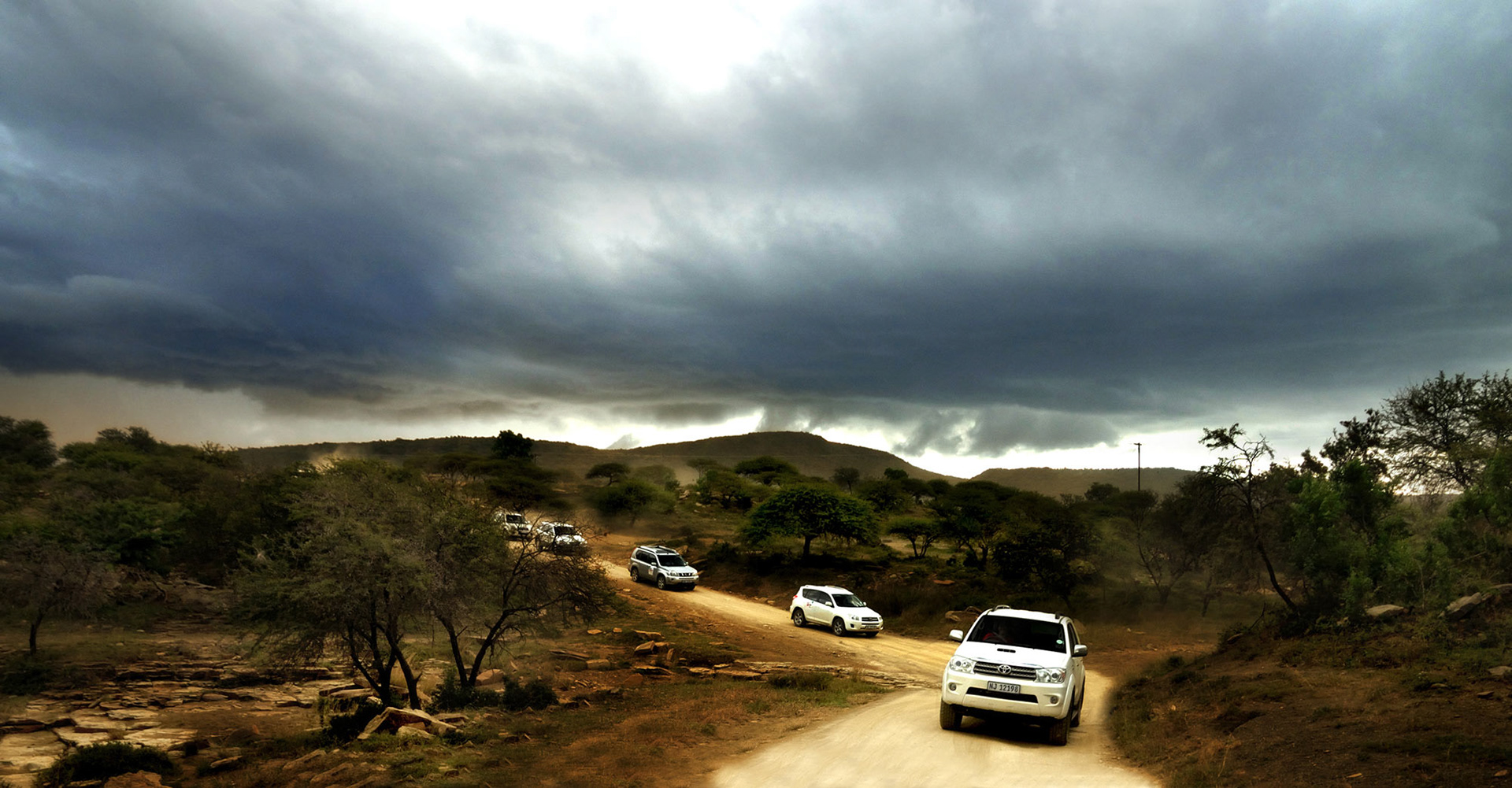 Convoy of white SUVs drives on a dirt road through a hilly landscape under a dramatic, stormy sky. Dark clouds and scattered trees enhance the sense of adventure.