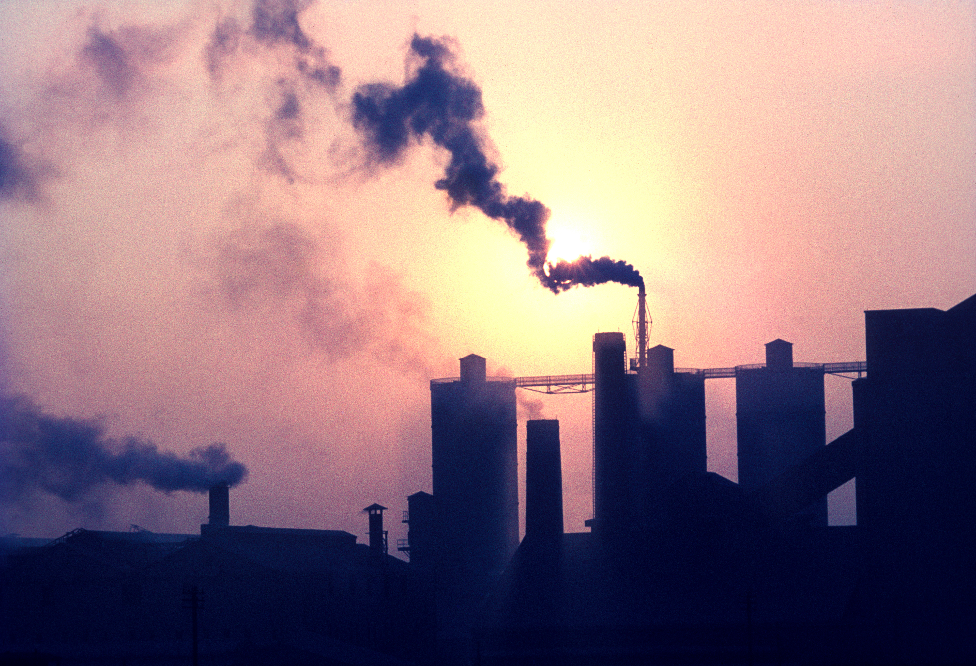 Silhouetted industrial chimneys emit dark smoke against a hazy, orange sunset sky, conveying pollution and industrial impact.