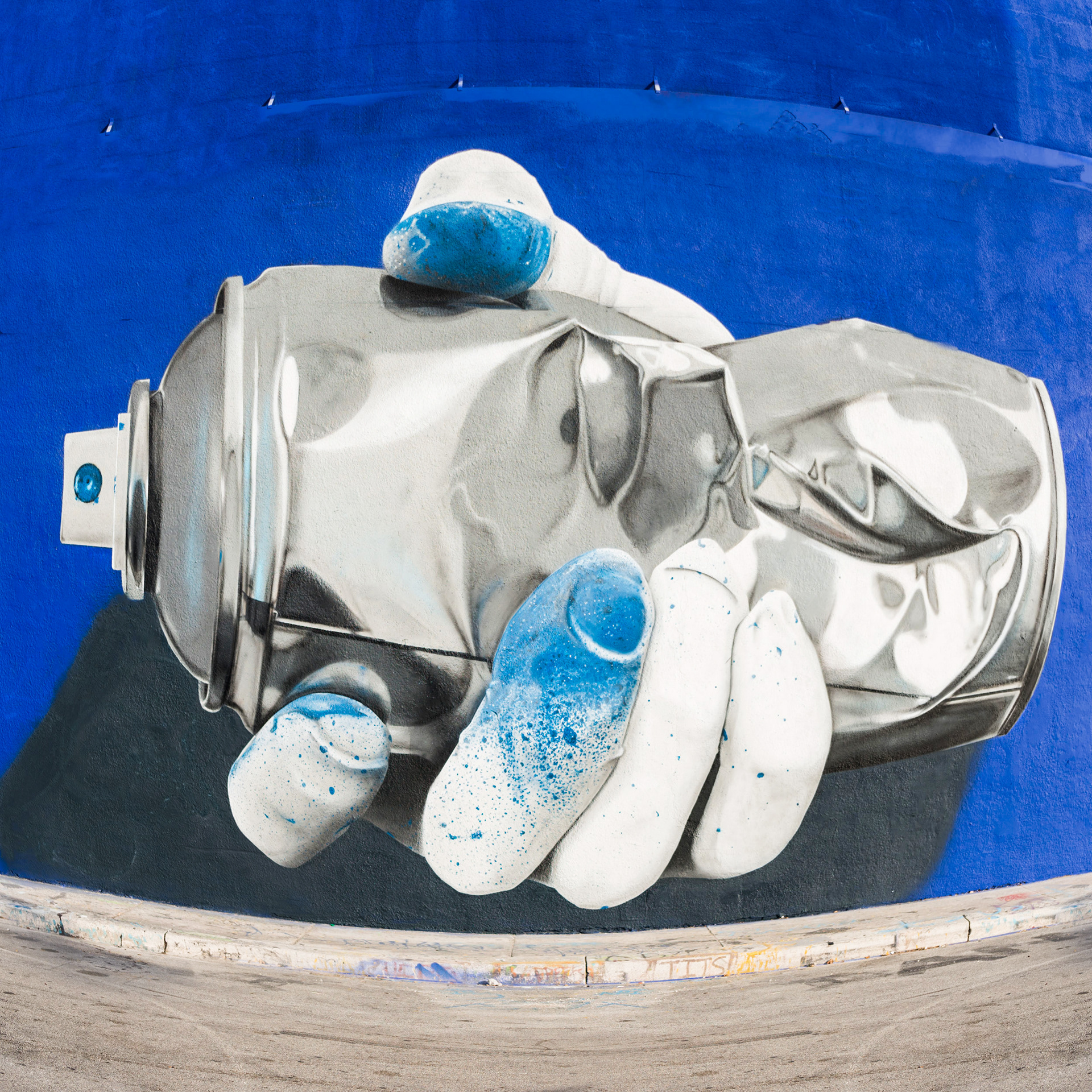 Street art mural of a hand holding a spray paint can against a vivid blue background. The hand is detailed with blue paint splatters, conveying creativity.