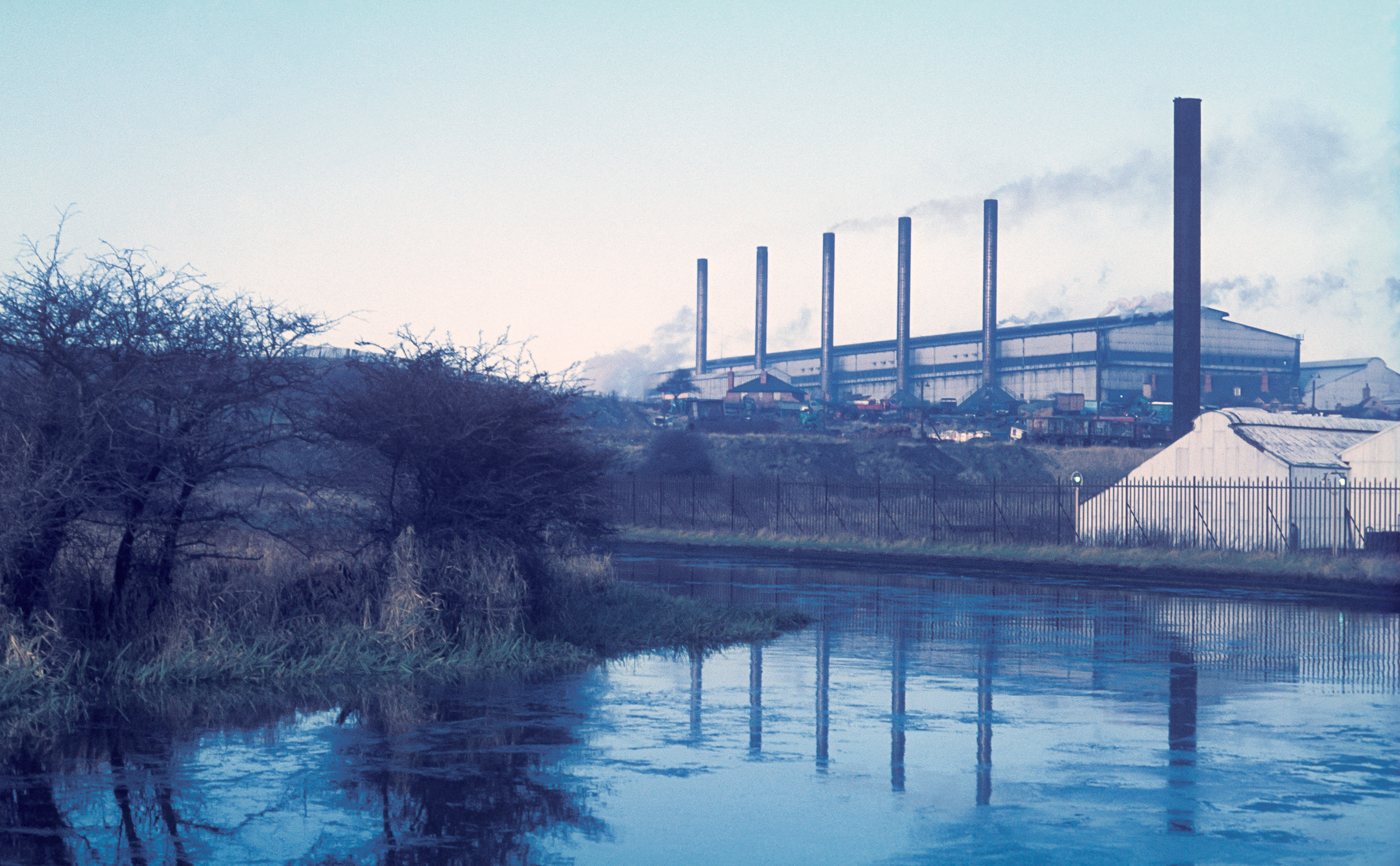 A factory Round Oak Steel Works with tall chimneys emits smoke, set against a pale blue sky. Bare trees and a calm river are in the foreground, creating a sombre, industrial scene now Merry Hill
