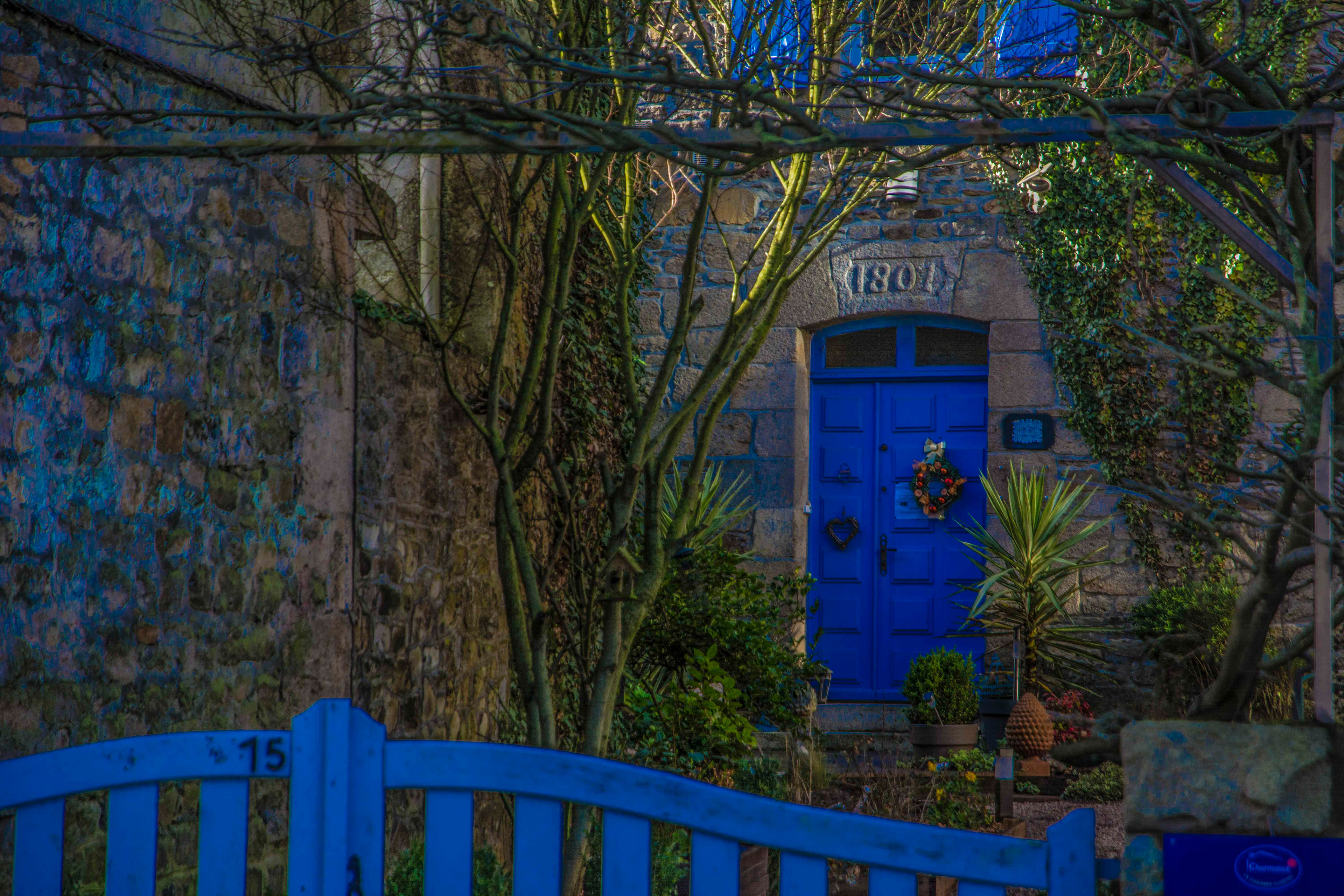 Stone house facade with a bright blue door, dated 1801. A festive wreath adorns the door. Leafy plants and ivy add a touch of nature and serenity.