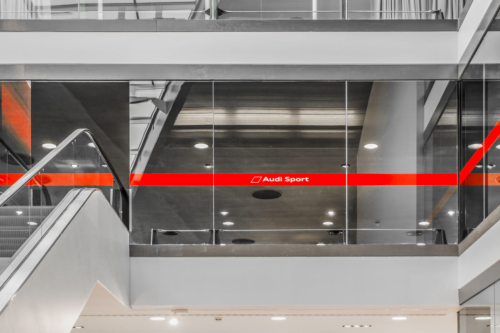 Modern interior with sleek glass panels and a bold red "Audi Sport" banner. Escalator on the left, ceiling lights reflecting on glossy surfaces.
