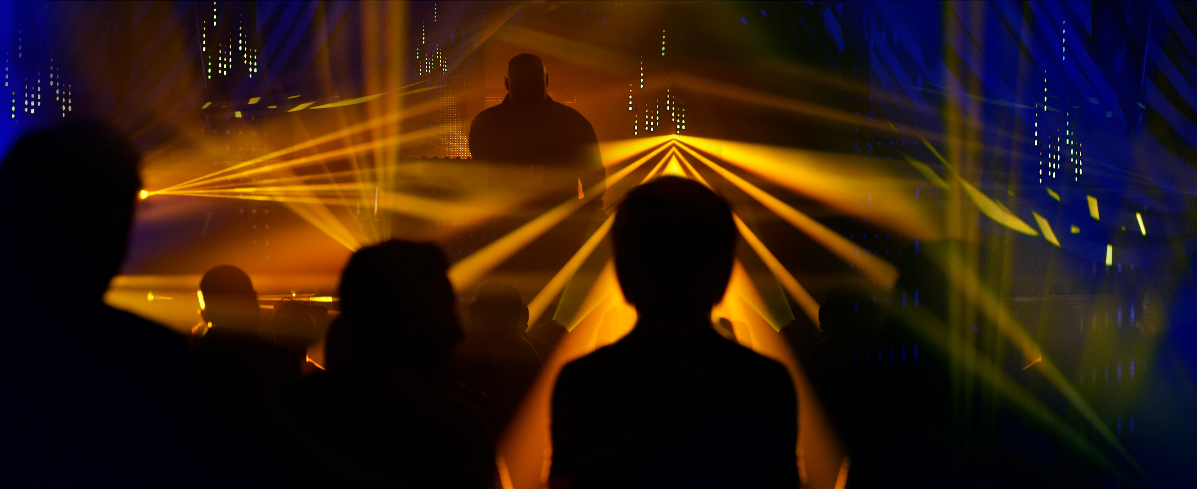 Silhouettes of people in a dark club with vibrant yellow laser lights and blue LED patterns. The atmosphere is lively and energetic.