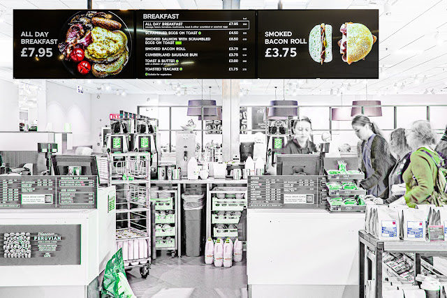 Checkout area of a café with digital menu boards overhead. Staff assist customers. Menu highlights all-day breakfast for £7.95, and offers a warm, inviting atmosphere.