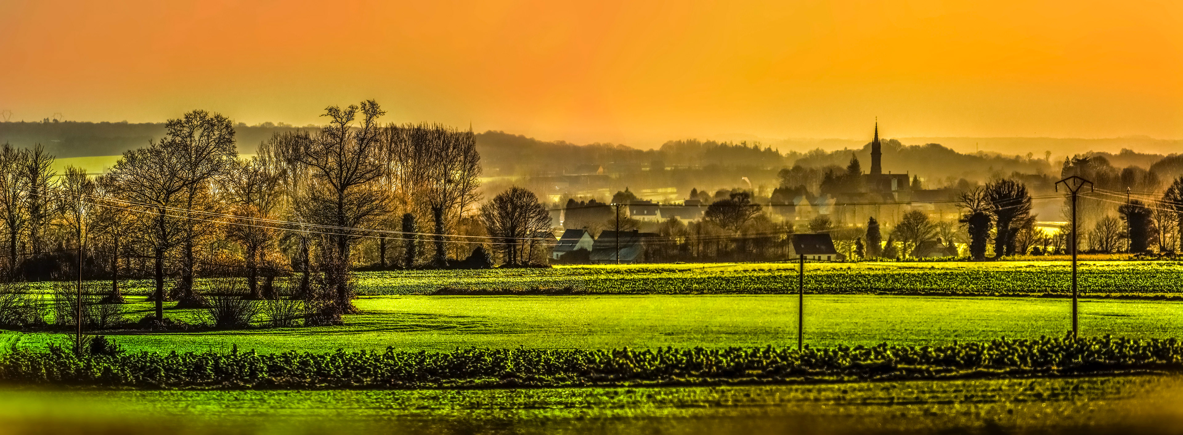 A serene rural landscape at sunset with silhouetted trees, green fields, and distant buildings under a vibrant orange sky, evoking tranquility.