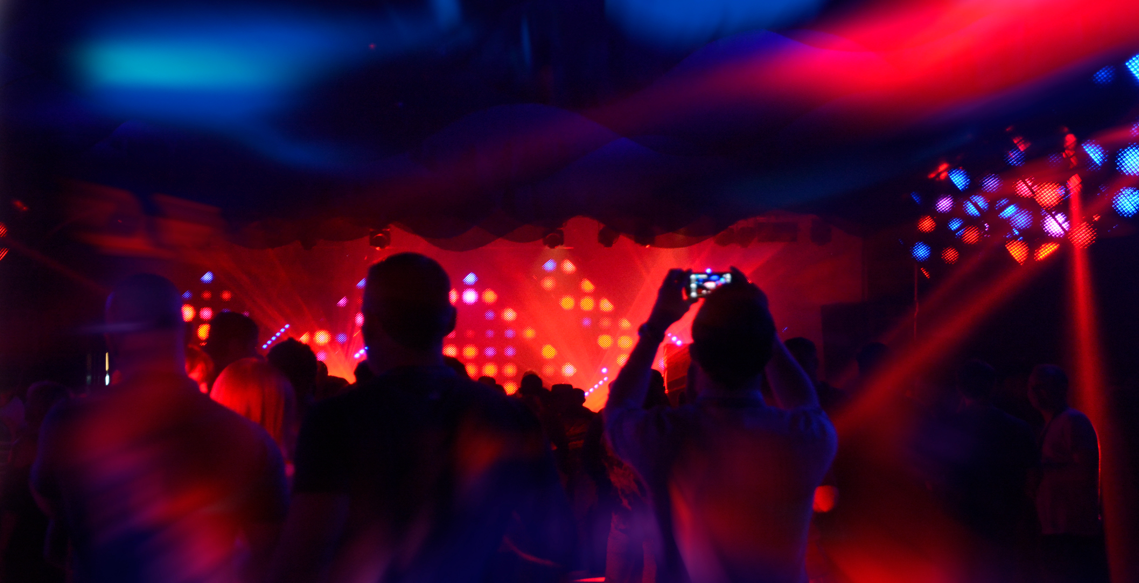 Silhouetted crowd at a concert, vibrant red and blue stage lights, person taking a photo with a phone, energetic and lively atmosphere.