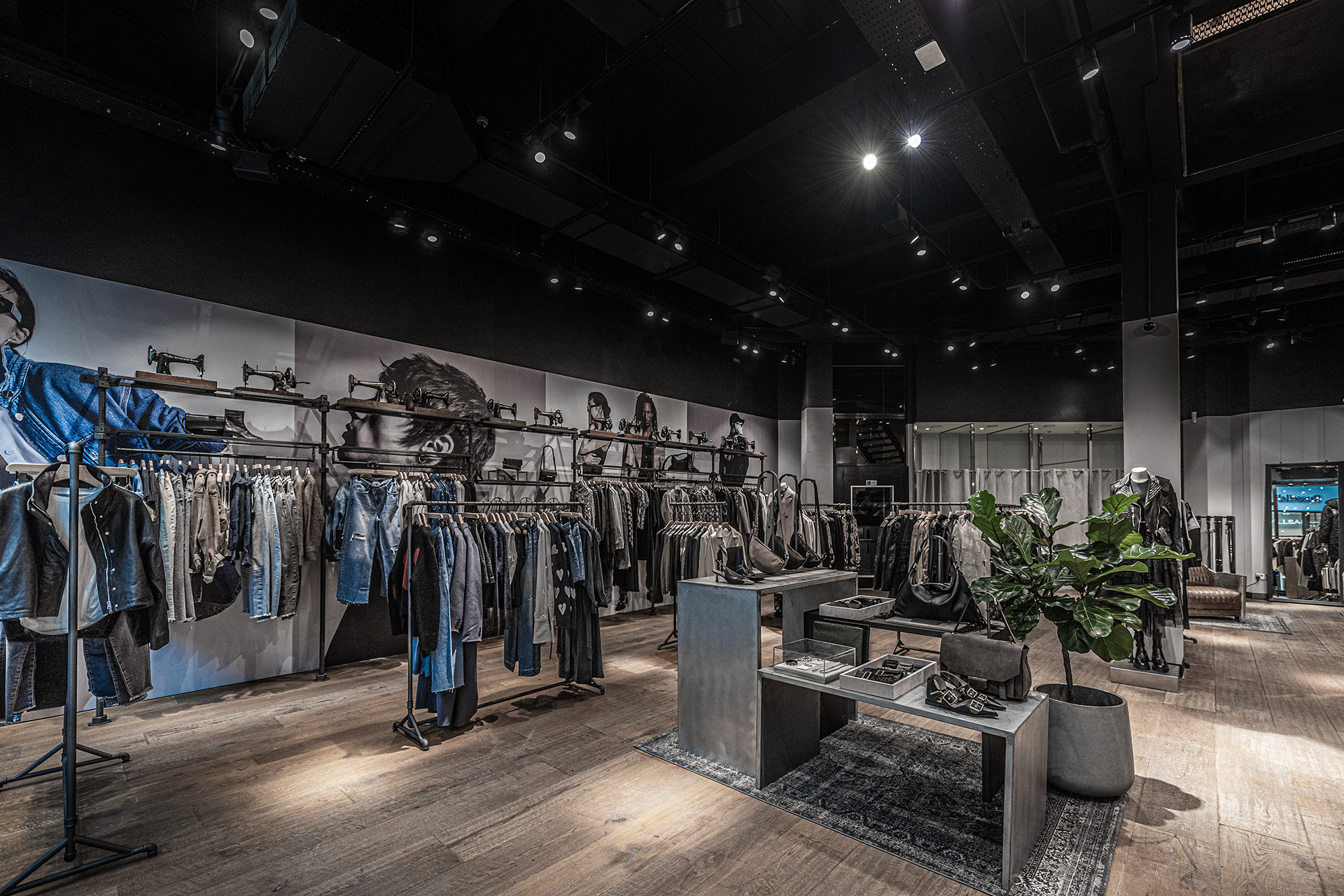 Trendy clothing store interior with dim lighting and modern decor. Racks display assorted denim and jackets under large wall posters. A potted plant adds greenery.