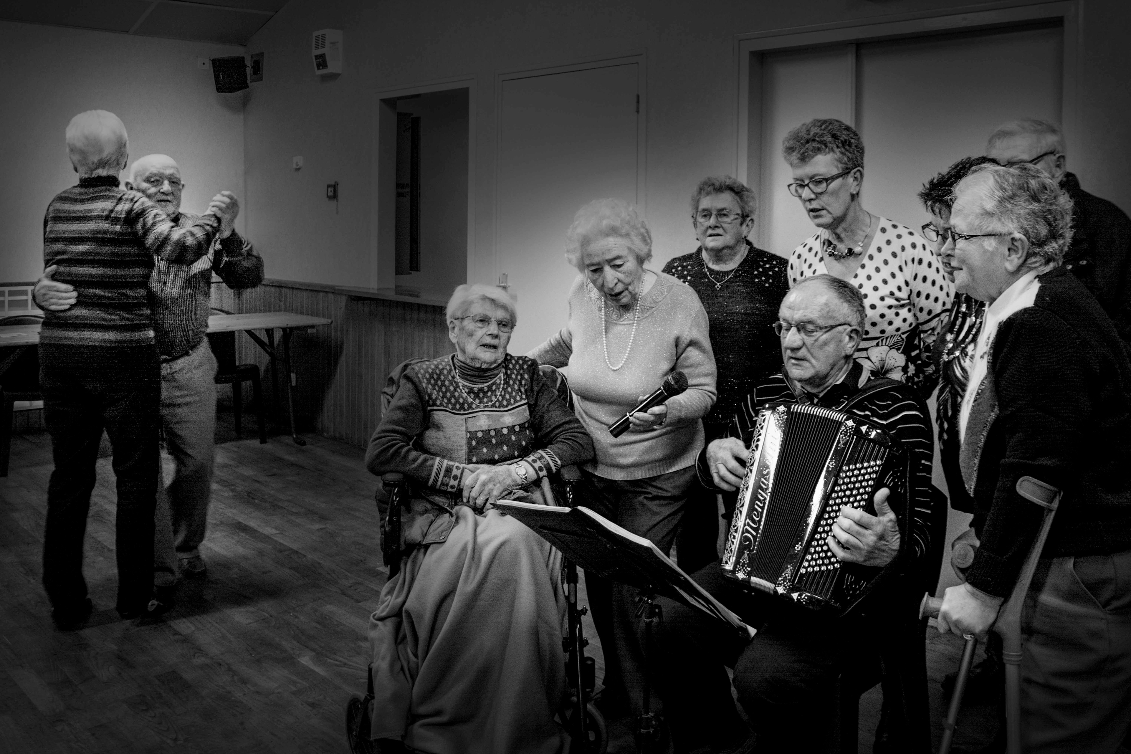 Elderly group in a room: a couple dances happily; others gather around a man playing an accordion, creating a warm, nostalgic atmosphere.