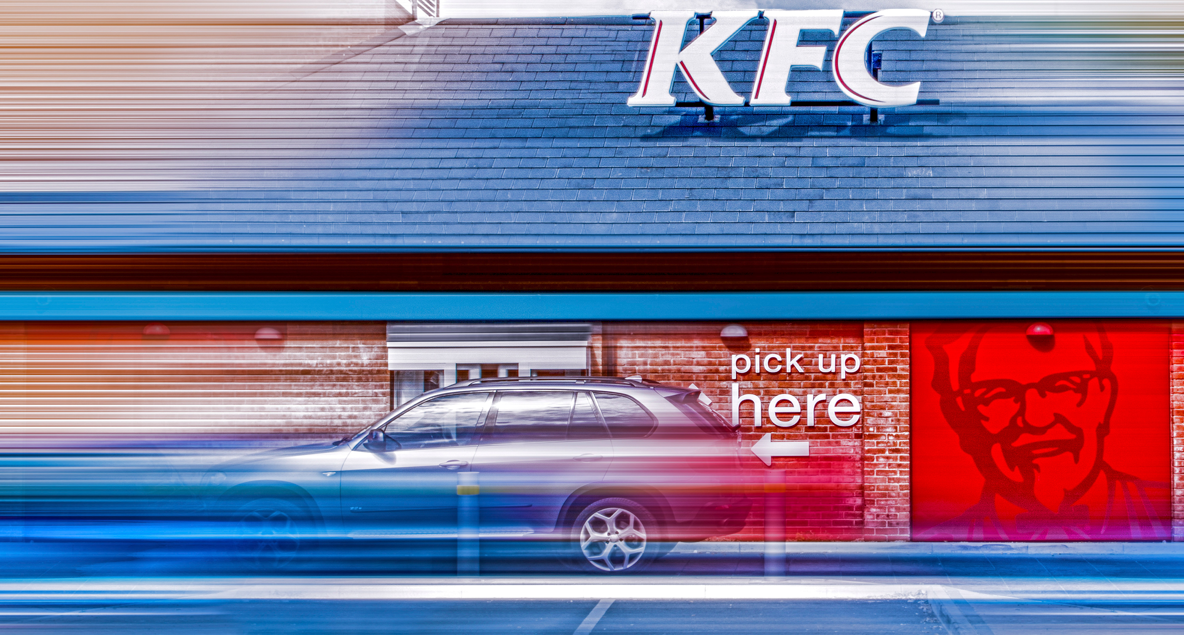 A blurred motion effect image of a KFC drive-thru with a dark SUV at the "pick up here" window. The Colonel's face is visible on the red wall