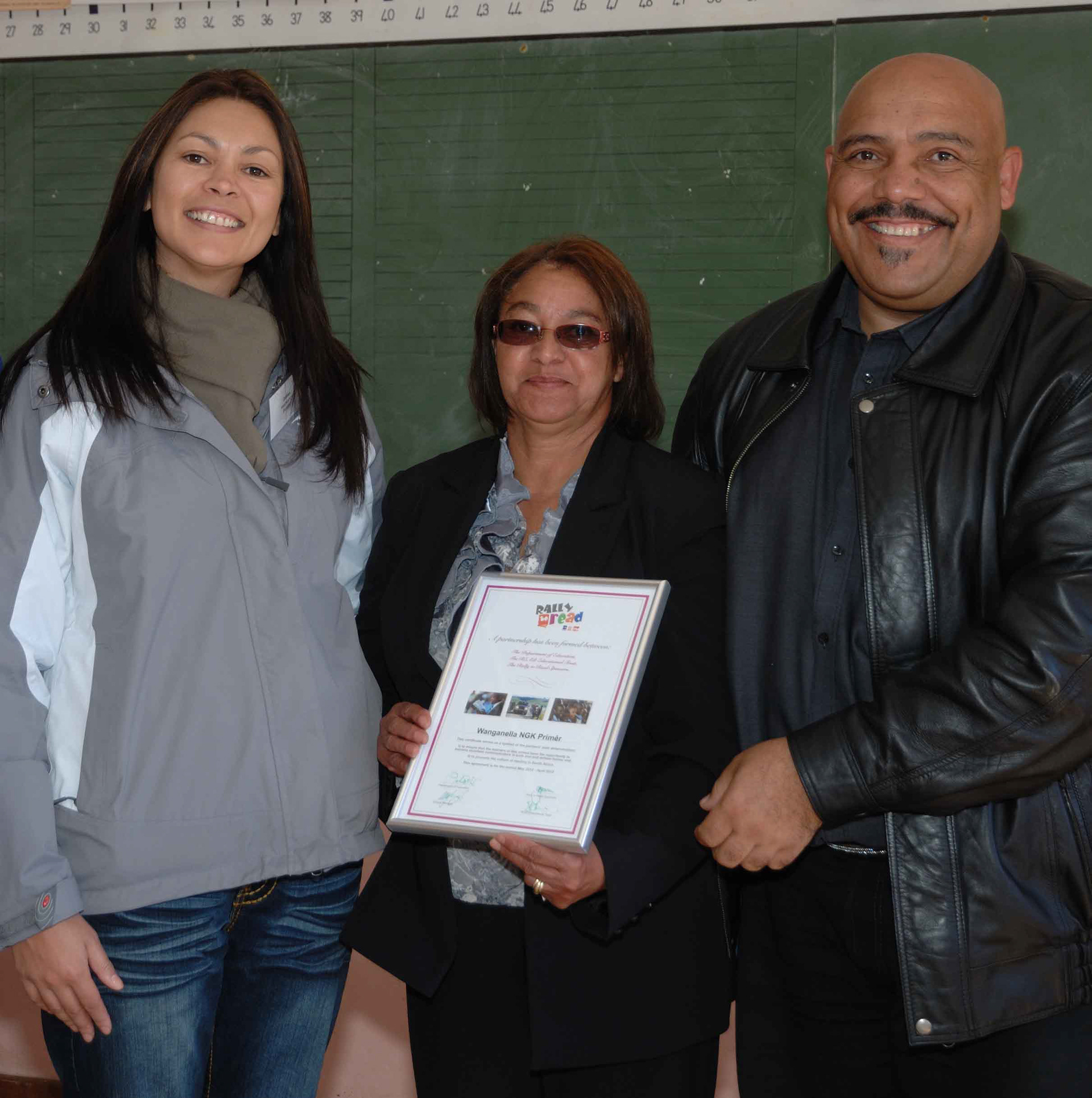 Three people smiling, with a middle-aged woman holding a framed certificate. They stand against a green wall, conveying a celebratory mood.