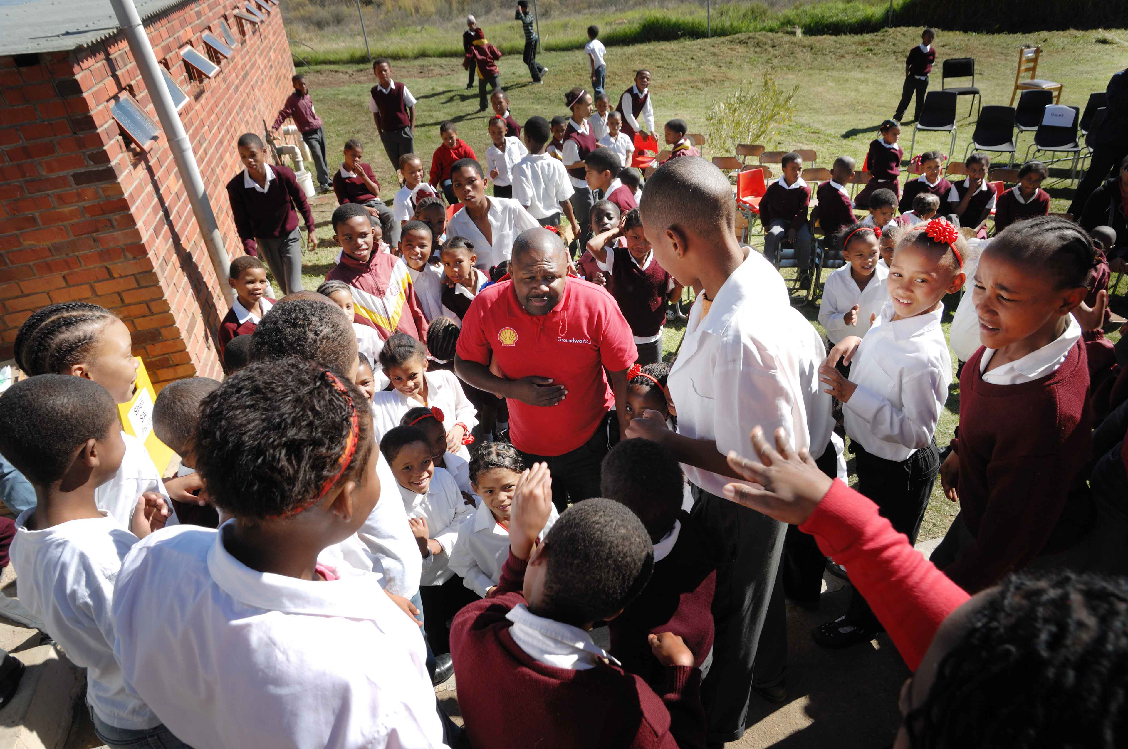 A group of school children in uniforms gather outside. An adult in a red shirt is speaking to them. The scene is lively and outdoor chairs are visible.