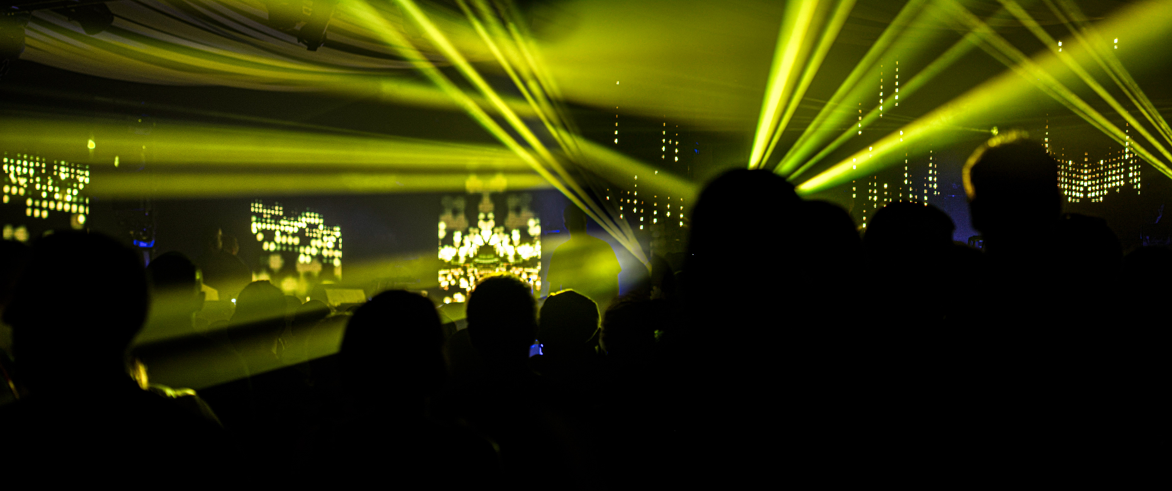 Silhouetted crowd at a concert, vibrant yellow laser beams illuminate the stage, creating an energetic and immersive atmosphere.