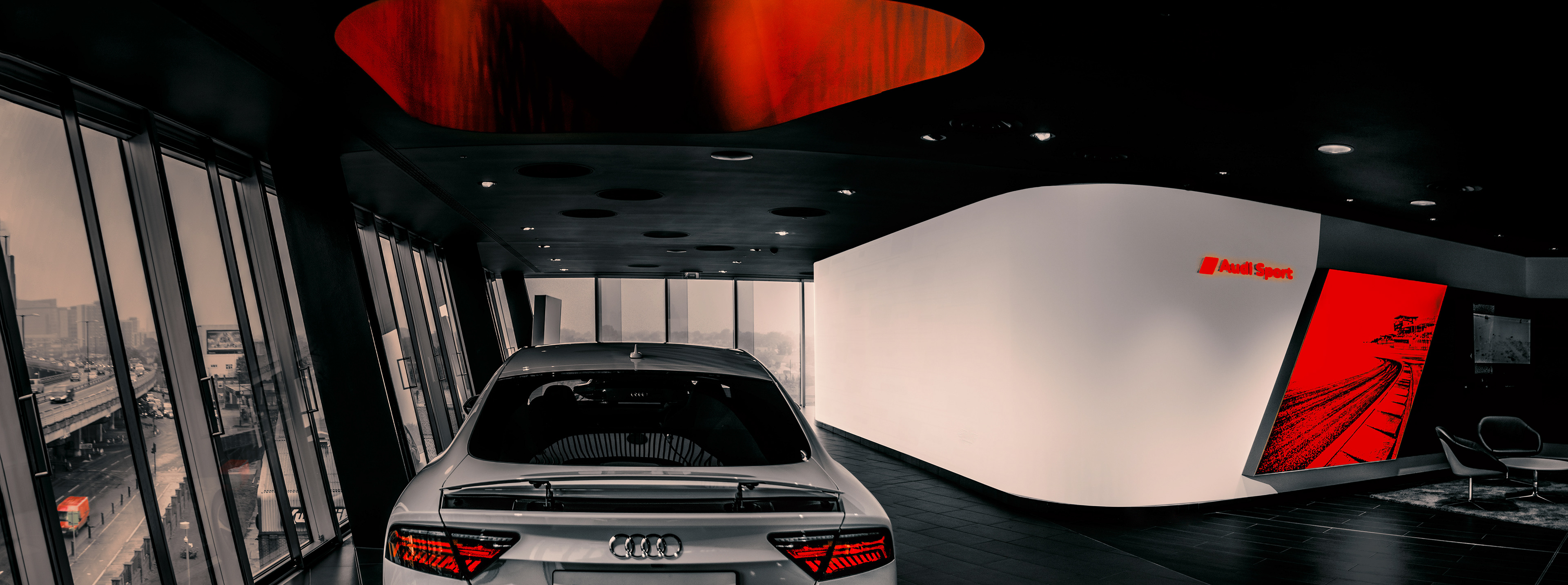 A sleek silver Audi car is in a modern showroom with large, slanted windows showing a cityscape outside. The interior is stylish, featuring a white wall with "Audi Sport" in red text and a vivid red display illustrating a racetrack. The atmosphere conveys luxury and innovation.