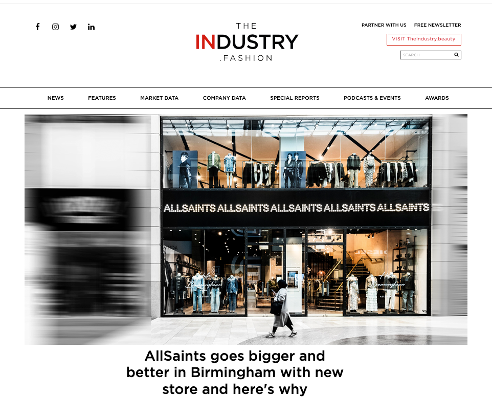 Storefront of AllSaints in Birmingham. Glass facade displays clothing, mannequins, and a glowing sign. A person walks by, creating a bustling city vibe.