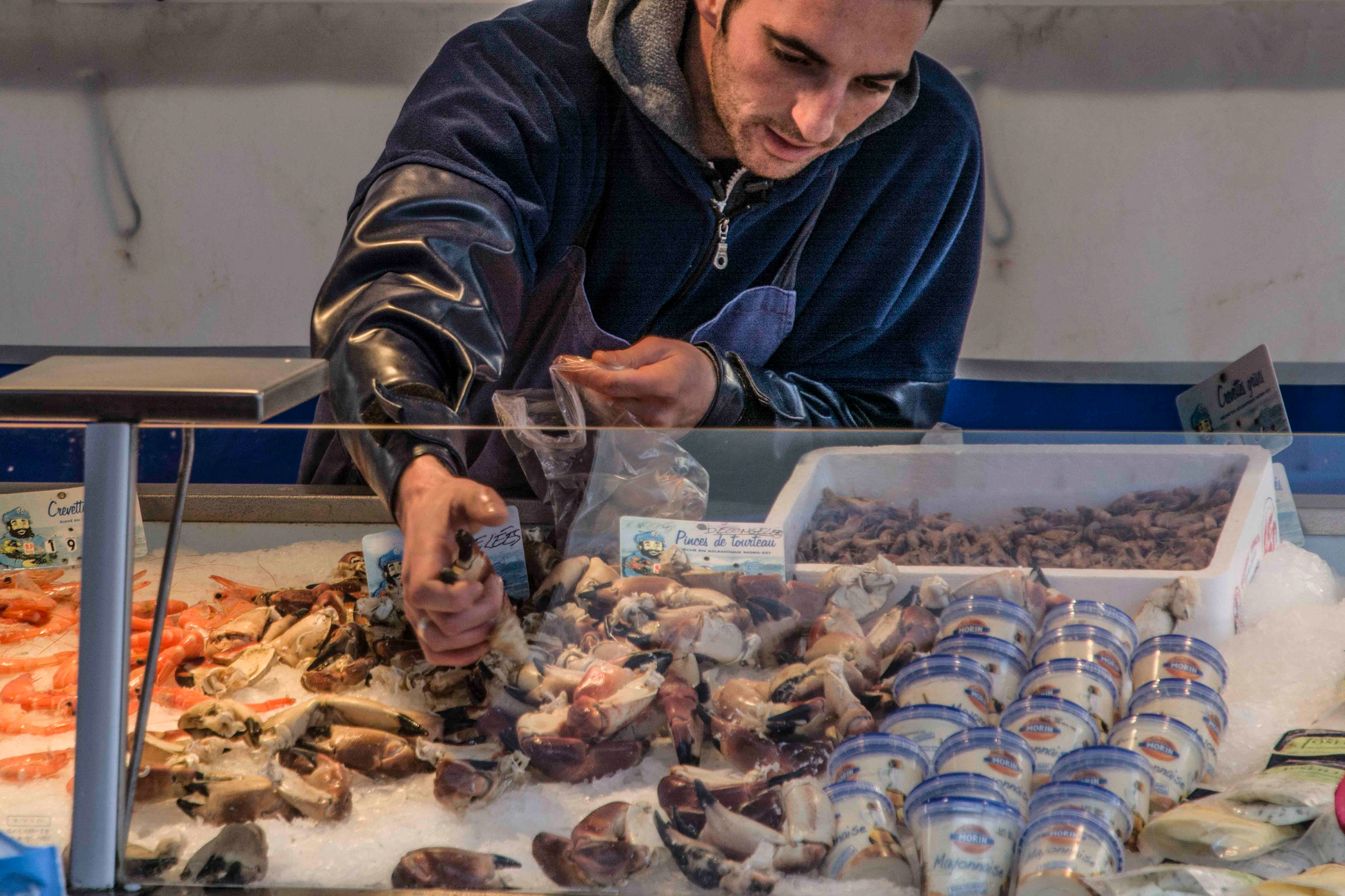A man in a dark jacket selects seafood from a market display filled with crab claws, shrimp, and sealed containers. The scene is busy and bustling market in rouillac france.