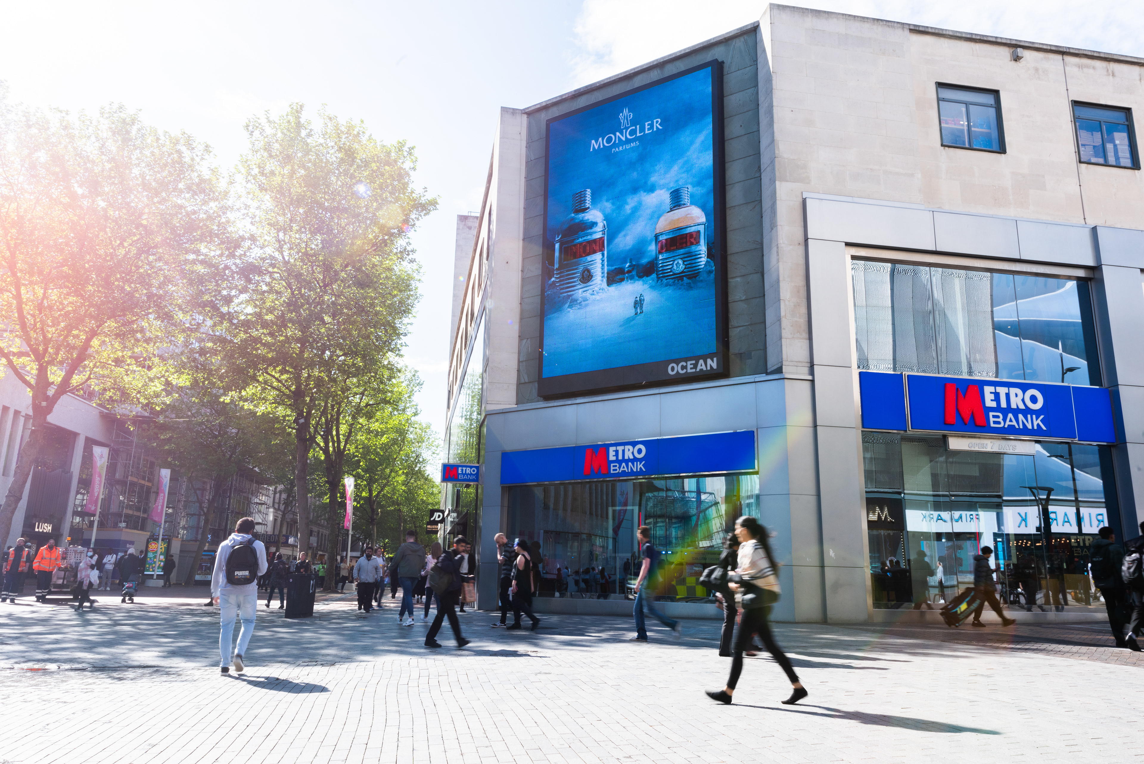 City scene with pedestrians in front of Metro Bank. Large Moncler advertisement on building, trees with sunlight in background, conveying a vibrant urban atmosphere. Ocean Outdoor photography