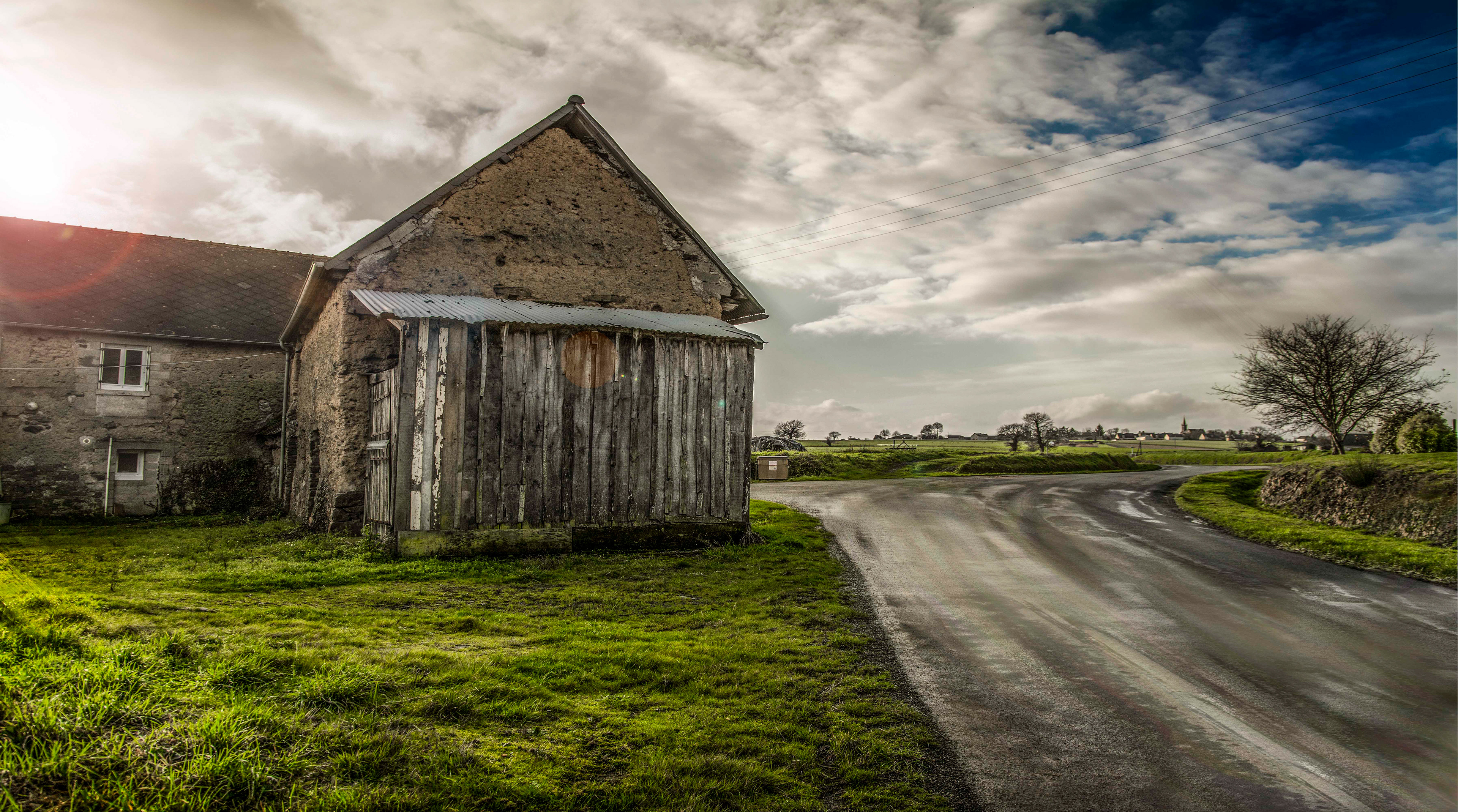 Rural scene with an old stone barn and wooden doors beside a curving road. Overcast sky and soft sunlight create a serene, nostalgic atmosphere.