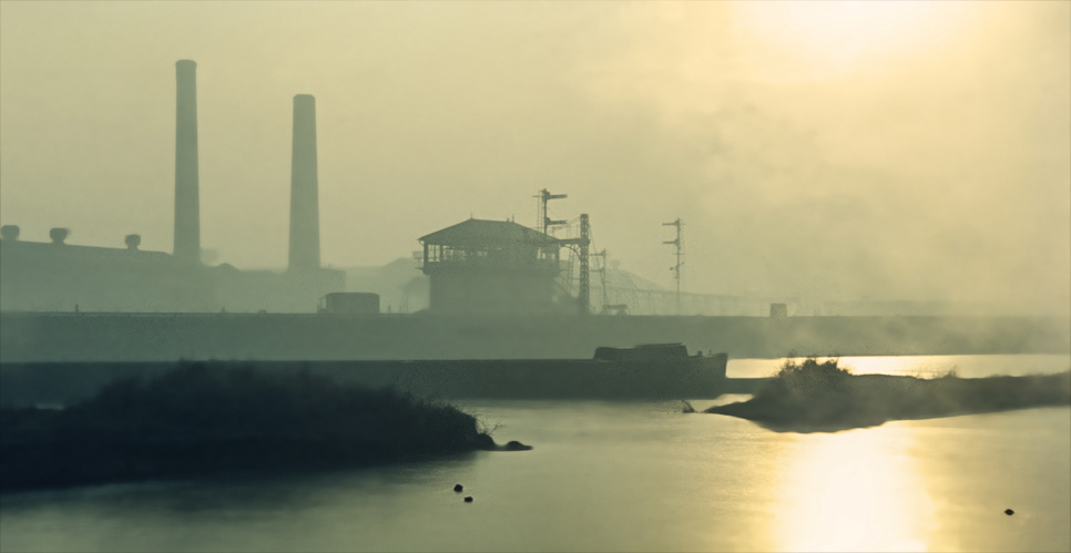 Foggy industrial landscape with dimly lit smokestacks, hazy structures, and a calm river reflecting soft light, evoking a tranquil yet sombre mood.