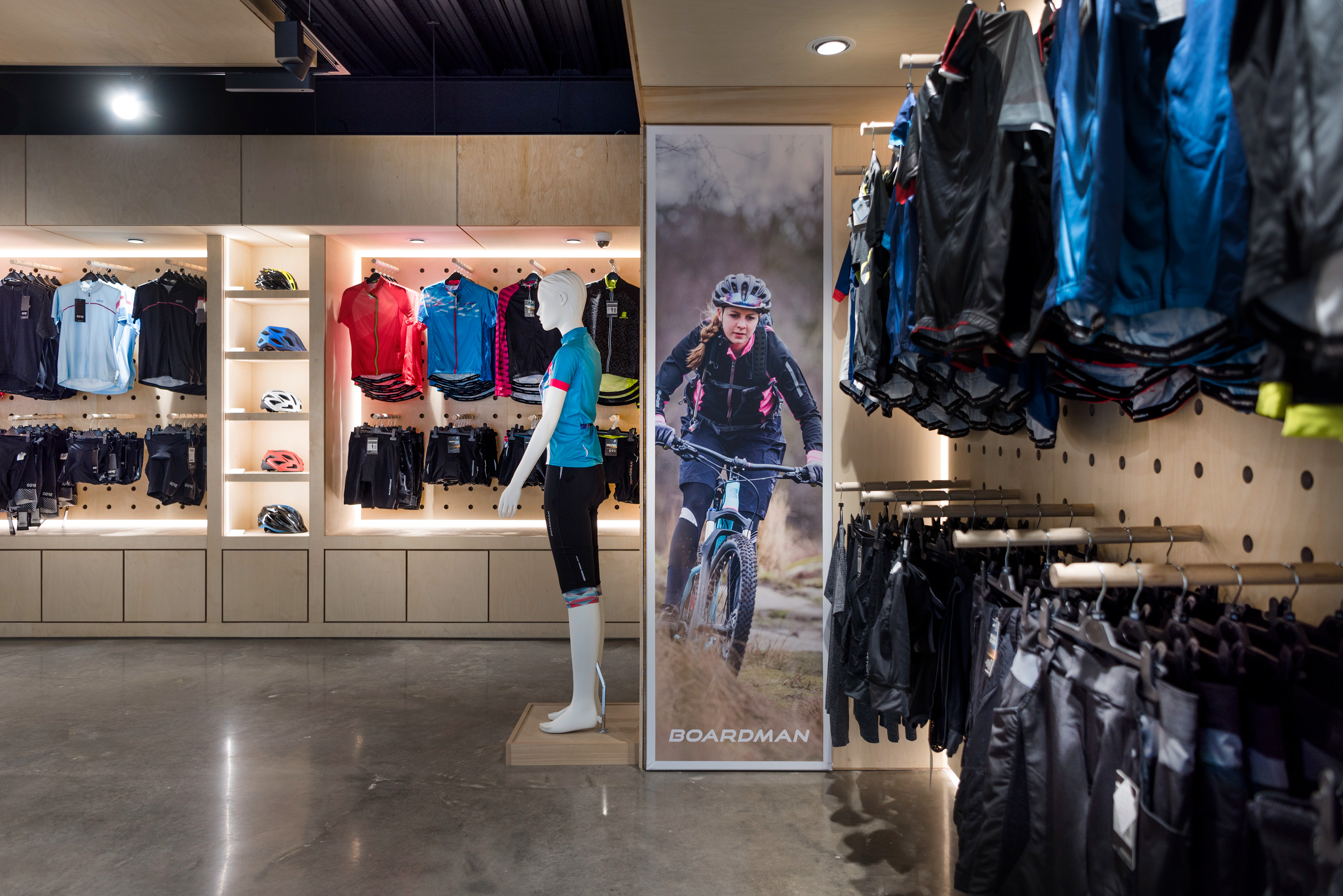 Store interior showcasing cycling apparel. Mannequin in cycling gear stands beside shelves of colourful jerseys and helmets. A cycling poster adds an adventurous tone.
