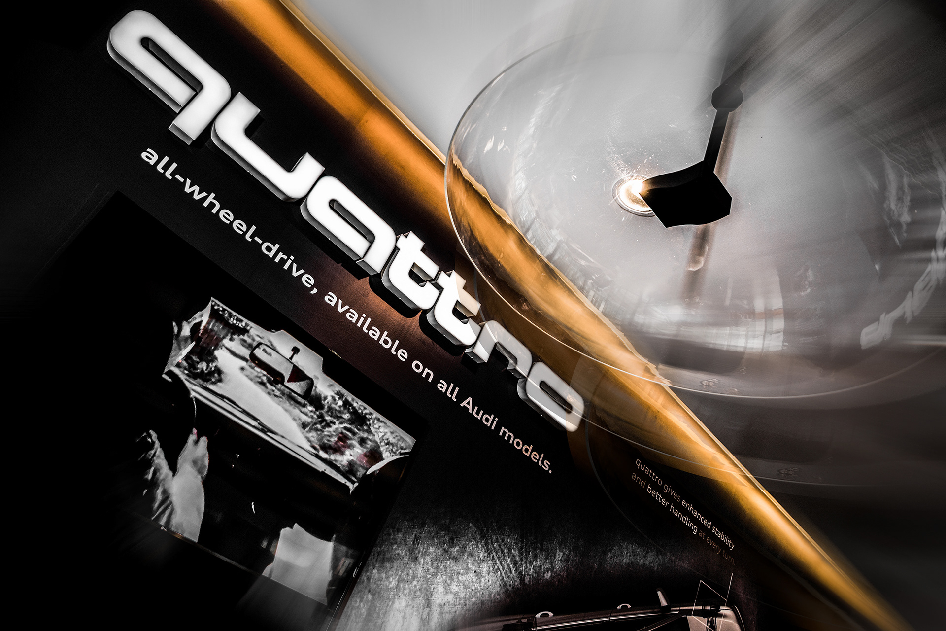 Bold "quattro" text in white, with "all-wheel-drive, available on all Audi models" below it. A blurred image evokes speed and technology.