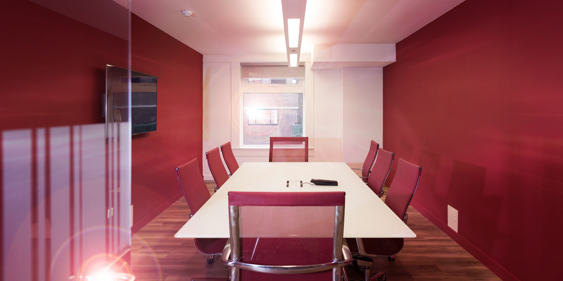 A modern conference room with red walls features a long white table and six red mesh chairs. A wall-mounted TV and window bring in natural light.