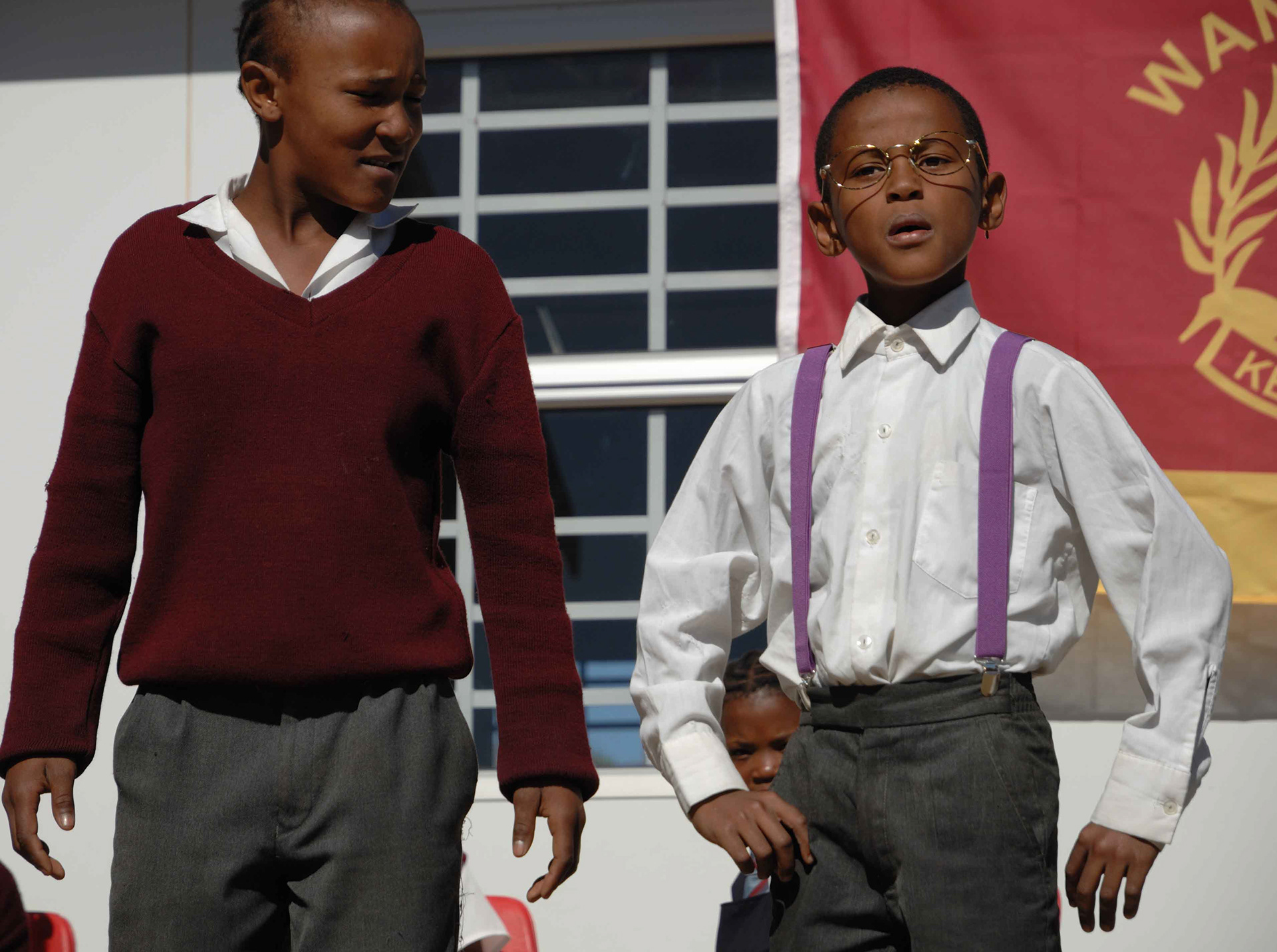 Two children stand outside in school uniforms. One wears a maroon sweater, the other a white shirt with purple suspenders. A red banner is behind them.