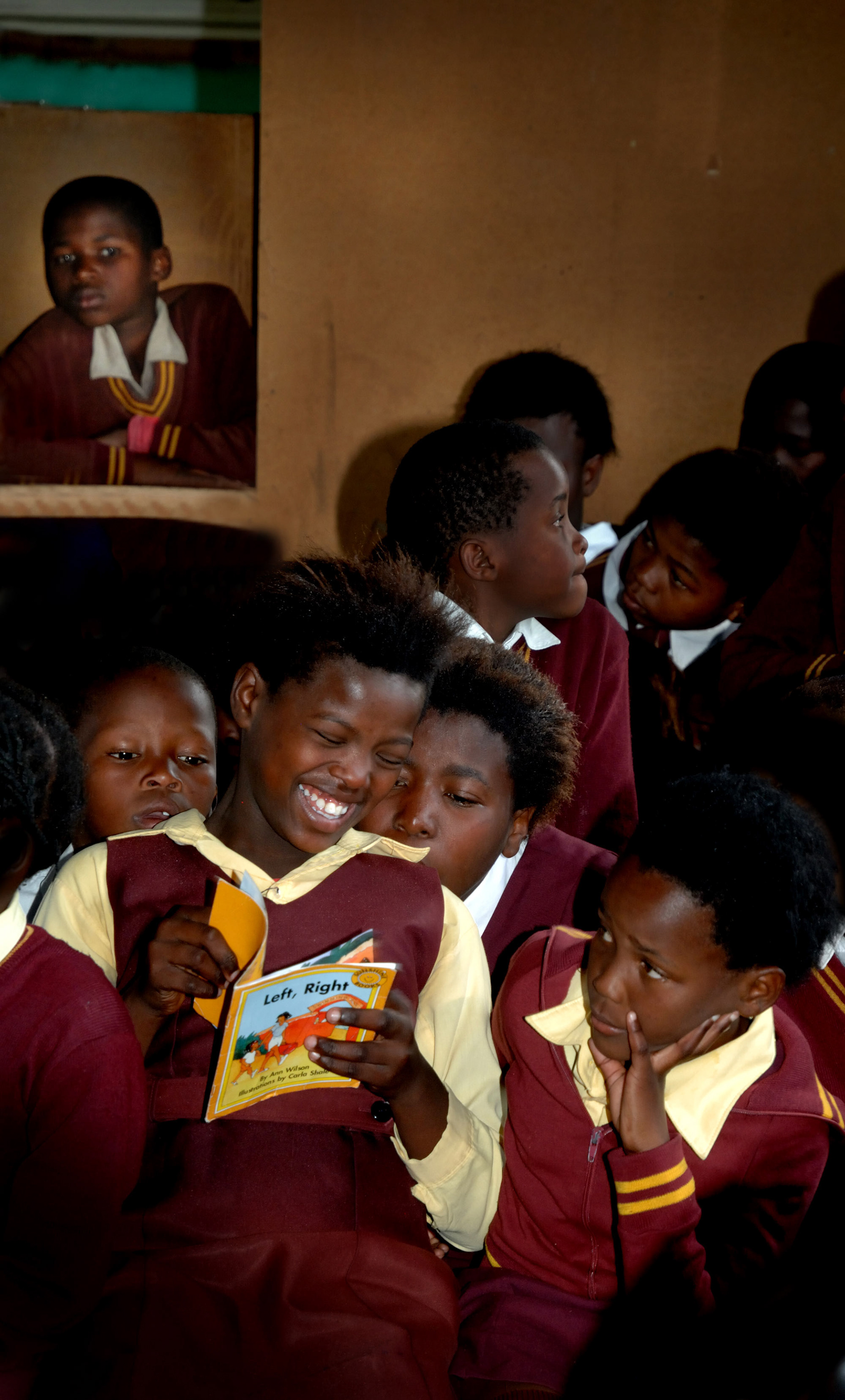 Children in school uniforms gather closely, sharing a book titled "Left, Right." One child smiles brightly, creating a joyful and engaged atmosphere.