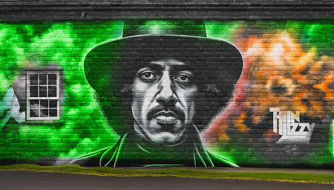 Street art mural of a man Phil Lynott in a wide-brimmed hat on a brick wall, surrounded by vibrant green, orange, and white swirls. "Thin Lizzy" is painted on the right.
