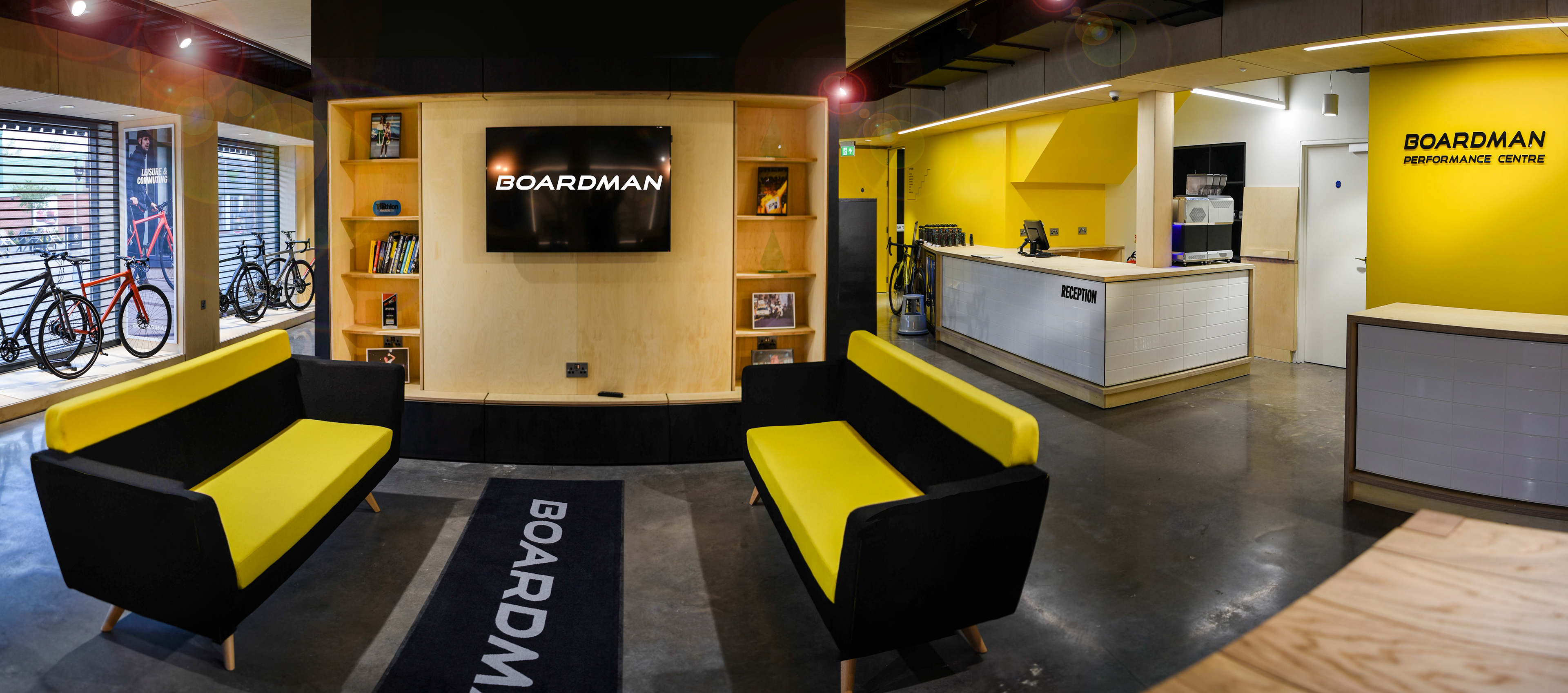 Modern showroom with yellow and black sofas, and a Boardman screen. Bicycles displayed by large windows, a sleek yellow reception area to the right.