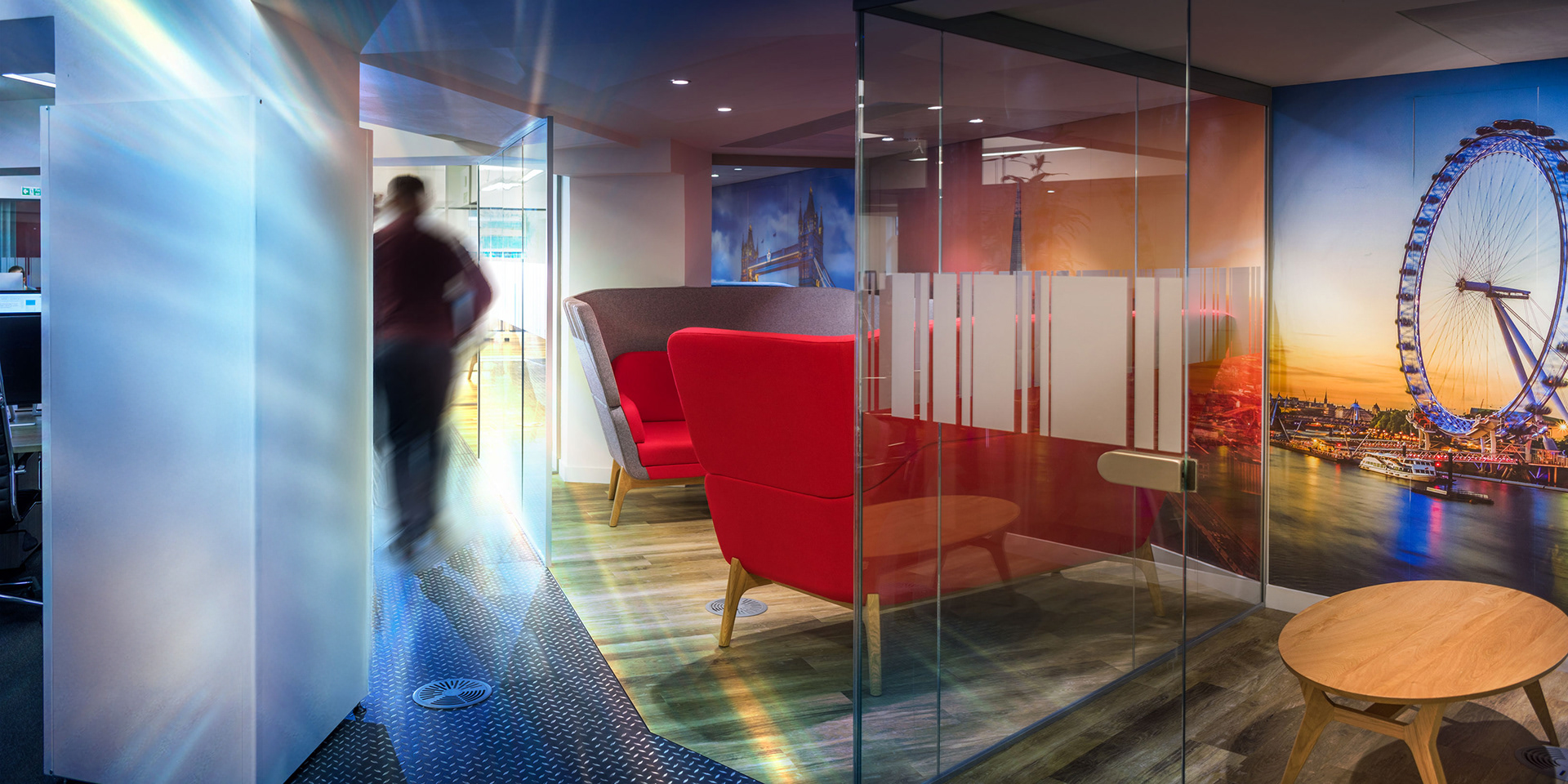 Modern office space with vibrant decor. Features red armchairs, a round wooden table, and a glass wall displaying a London Eye mural. A person walks by, creating motion blur.